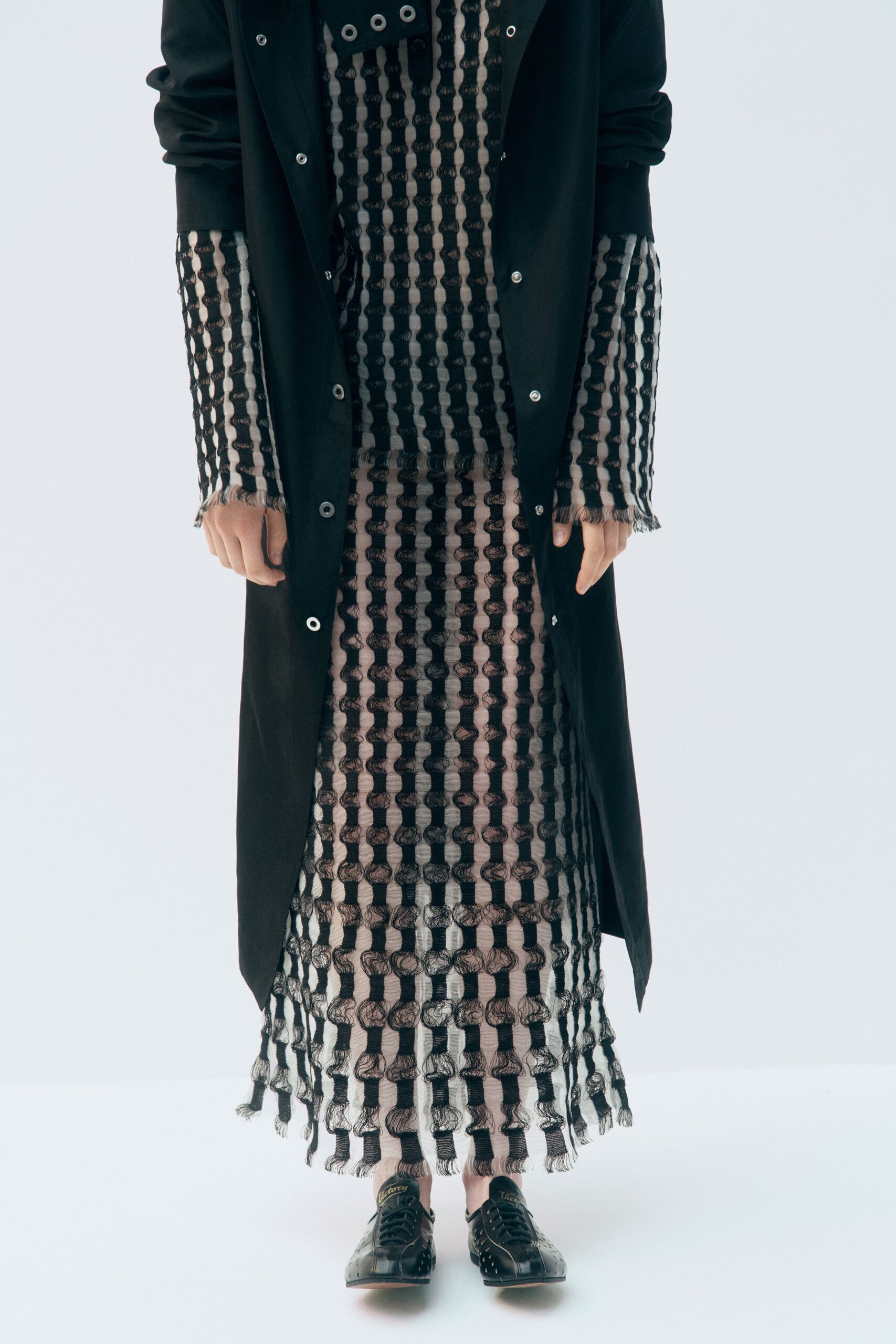 Black snap-front coat, silver grommets marching down the placket. Nude column dress latticed with black looped stripes and frayed cuffs. Black leather lace-ups punched with tiny metal-rim eyelets. Crossing a museum atrium while the rain still dots the stone steps.