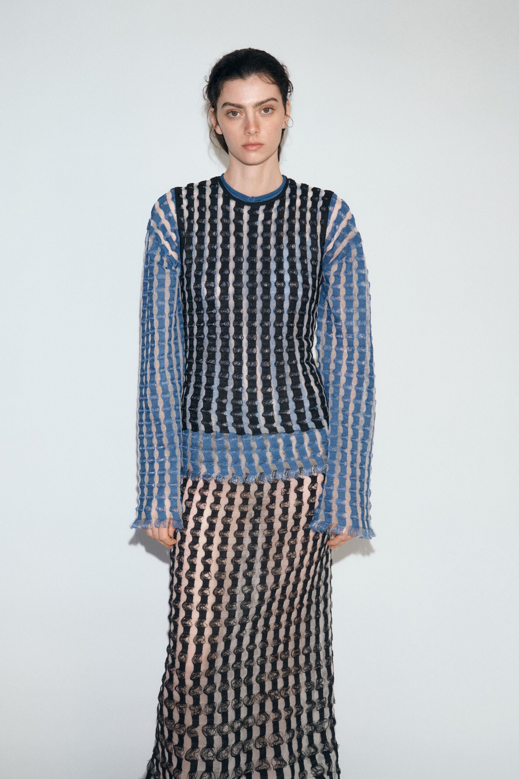 Black openwork mesh top with puckered vertical ribs over nude lining. Powder-blue long-sleeve mesh dress, frayed cuffs and hem skimming below. Column skirt in matching black openwork, the looped texture slightly skewed. Museum lobby after rain, still carrying the chill from outside.