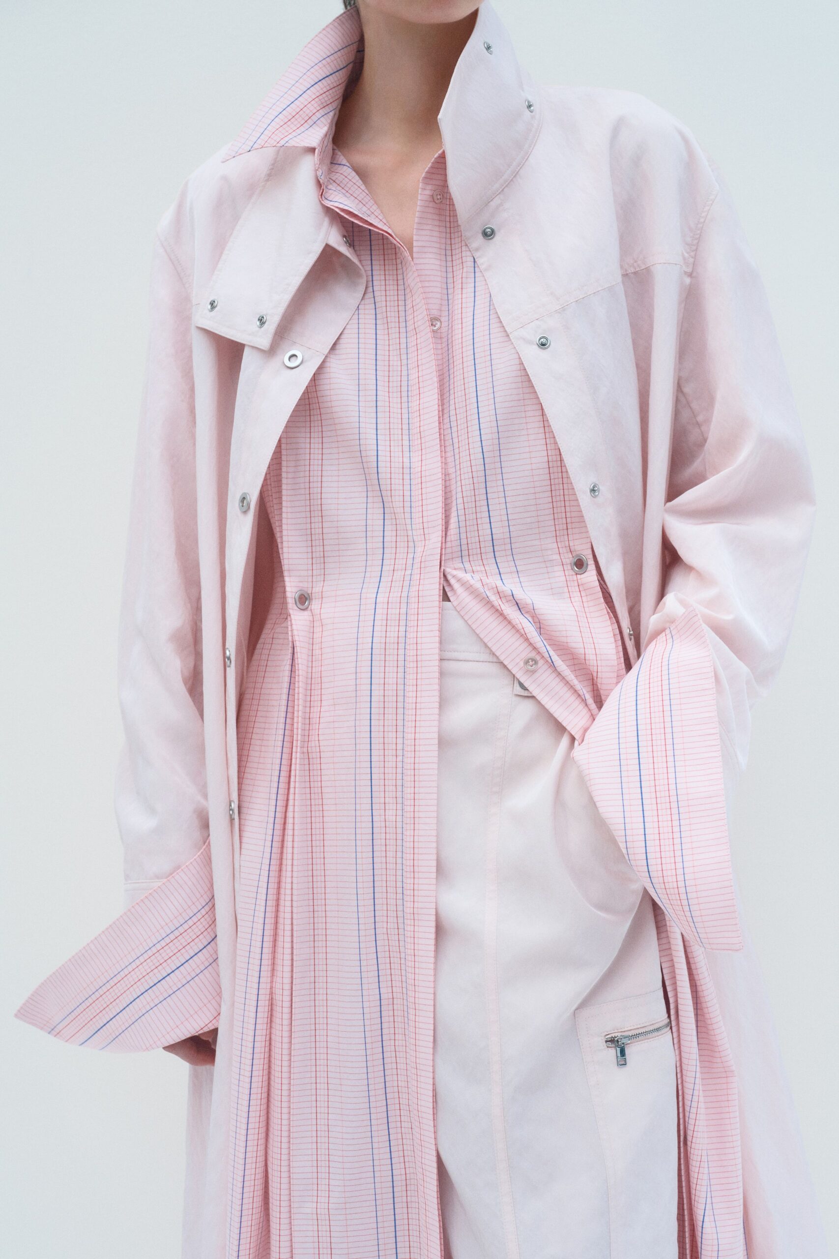 Powder-pink coat in crisp matte cloth, silver snaps climbing the stand collar. Blush graph-paper shirt with blue pinstripes and exaggerated turned-back cuffs. Pale blush trousers, one patch pocket finished with a small exposed zip. For the hour after a spring meeting when the office windows finally open.