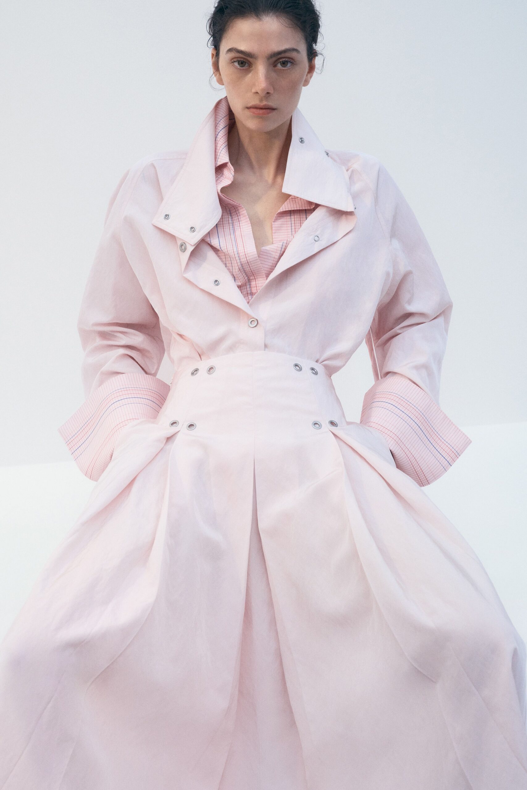 Pink graph-check shirt, collar loosened open beneath the outer layer. Pale blush cotton dress coat with snapped storm flap and exaggerated cuffs. Matching dome skirt, grommeted waistband and deep sculpted side folds. Wedding morning in a rented house, still in the sheets' pale light.