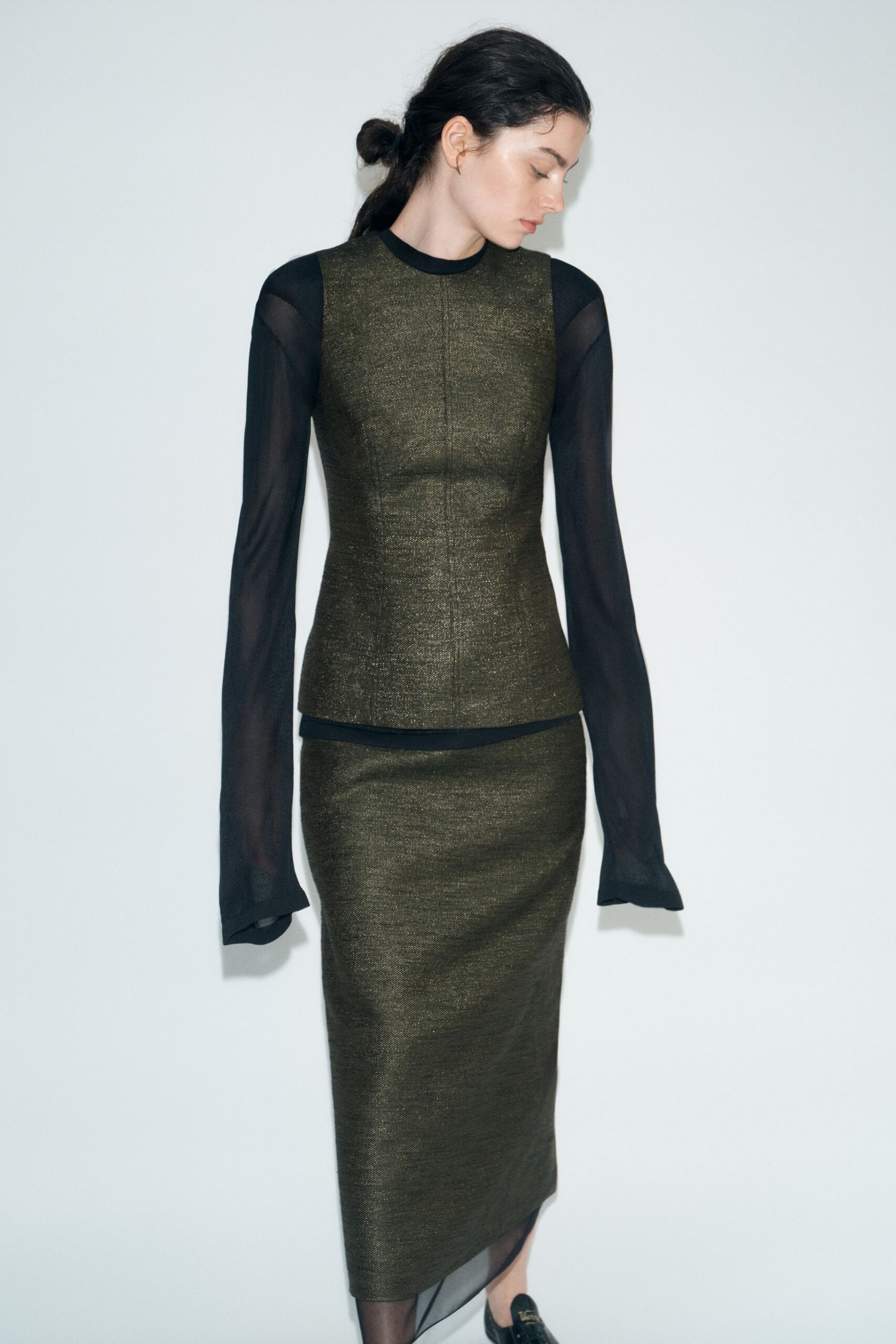 Olive metallic knit shell, center seam pulling slightly at the waist. Black sheer jersey sleeves with long cuffs drooping past the wrists. Matching pencil skirt in the same dull-shine knit. Black sheer tights. Black loafers with a small gold bit. For the hour before a museum benefit, alone in the service corridor.