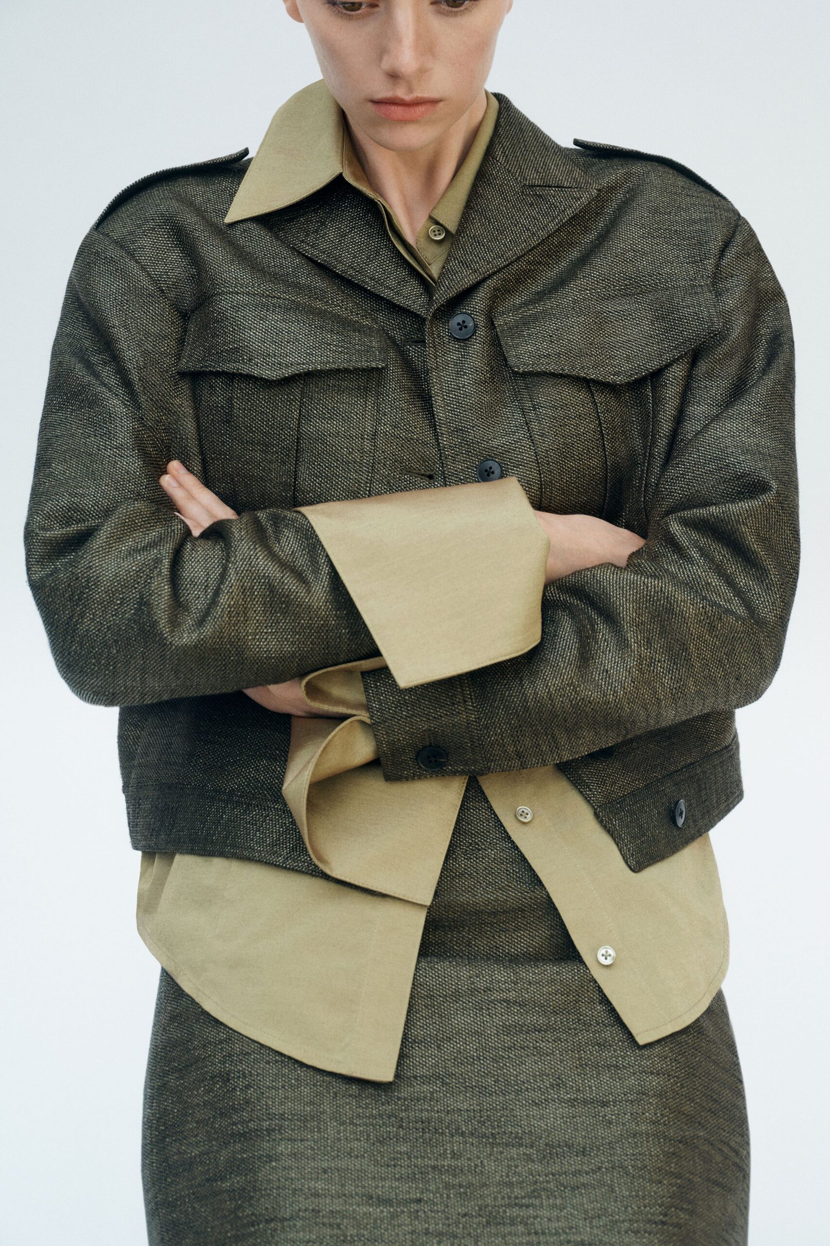 Boxy charcoal jacket in coarse basket-weave, flap yoke stitched across the chest. Sand shirt with exaggerated cuffs and collar, one small button peeking below. Matching straight skirt in the same dry woven cloth. For the kind of office where the radiators knock before dawn.