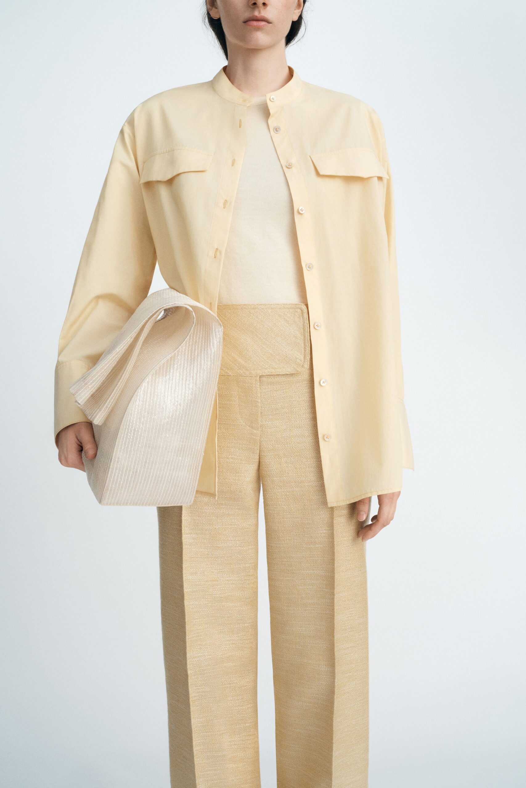 Buttercream overshirt with a band collar and flap chest panels. Fine-gauge cream knit shell, cut close through the torso. High-waisted oat trousers in dry basket-weave, front crease sharp. Large ivory pleated clutch, folded like a paper fan. Walking into a hotel breakfast room before the tables are reset.