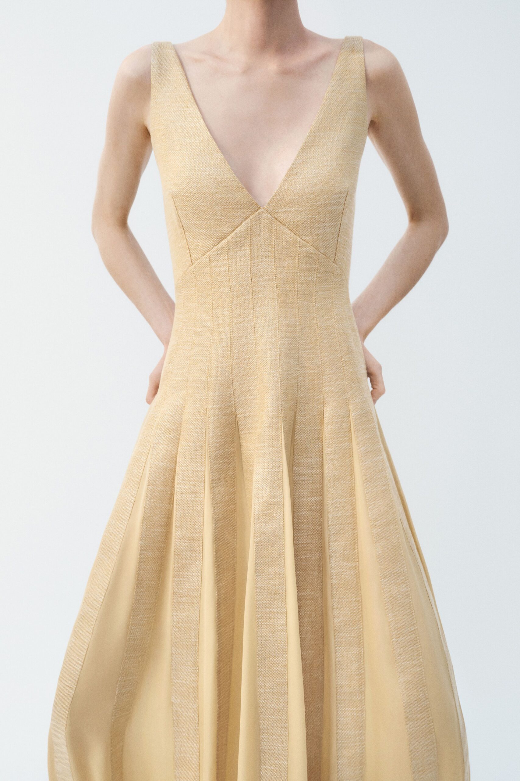 Buttercream dress in nubby woven bands over a plunging V, pleats opening soft between the panels. Afternoon wedding in a glasshouse, one heel sinking slightly into the lawn.