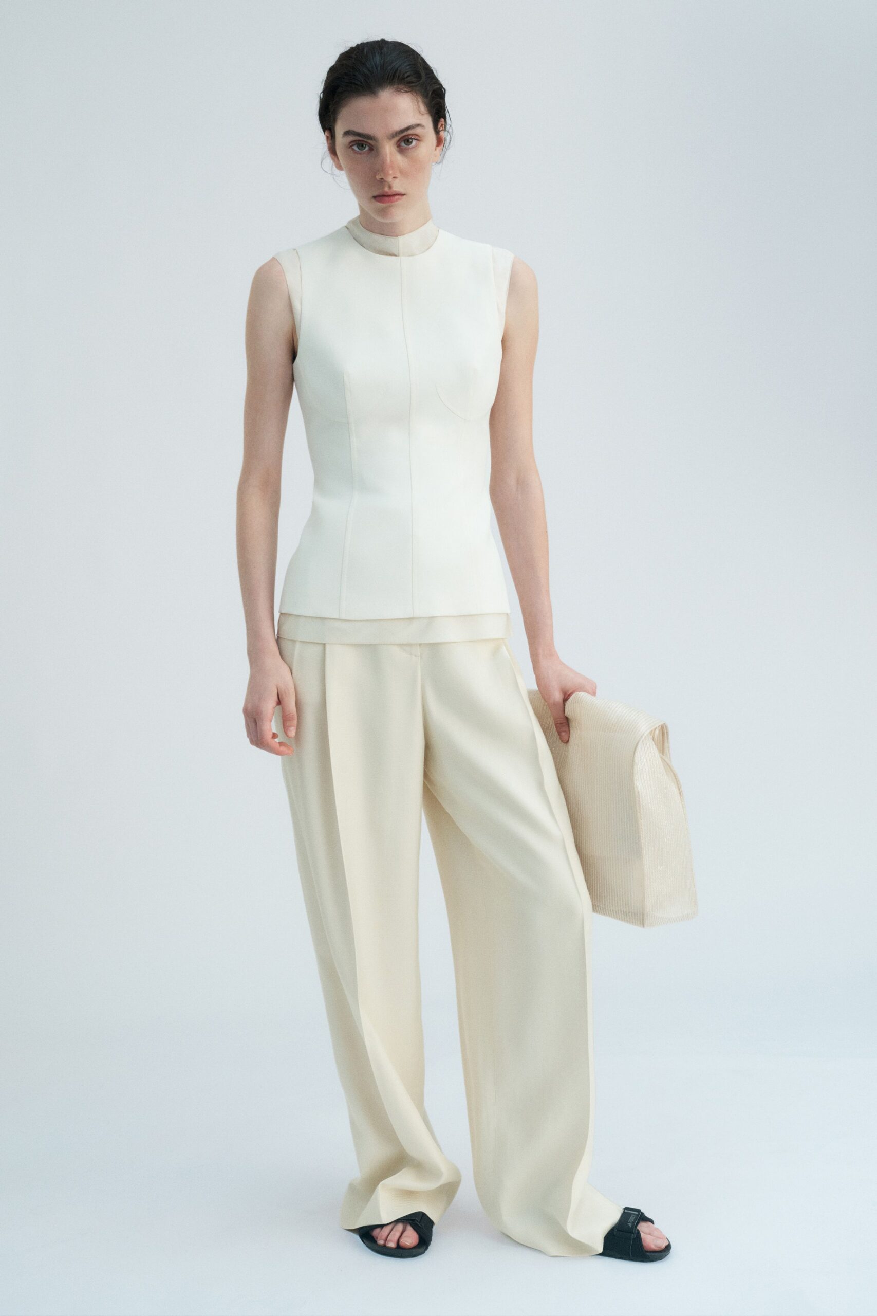 Cream sleeveless shell in dense suiting, one curved bust seam visible. Wide cream trousers with deep front pleats and pooled hems. Large ribbed ivory tote, tucked underhand like a folded panel. Black slide sandals with broad padded straps. For crossing a silent lobby before anyone else checks in.