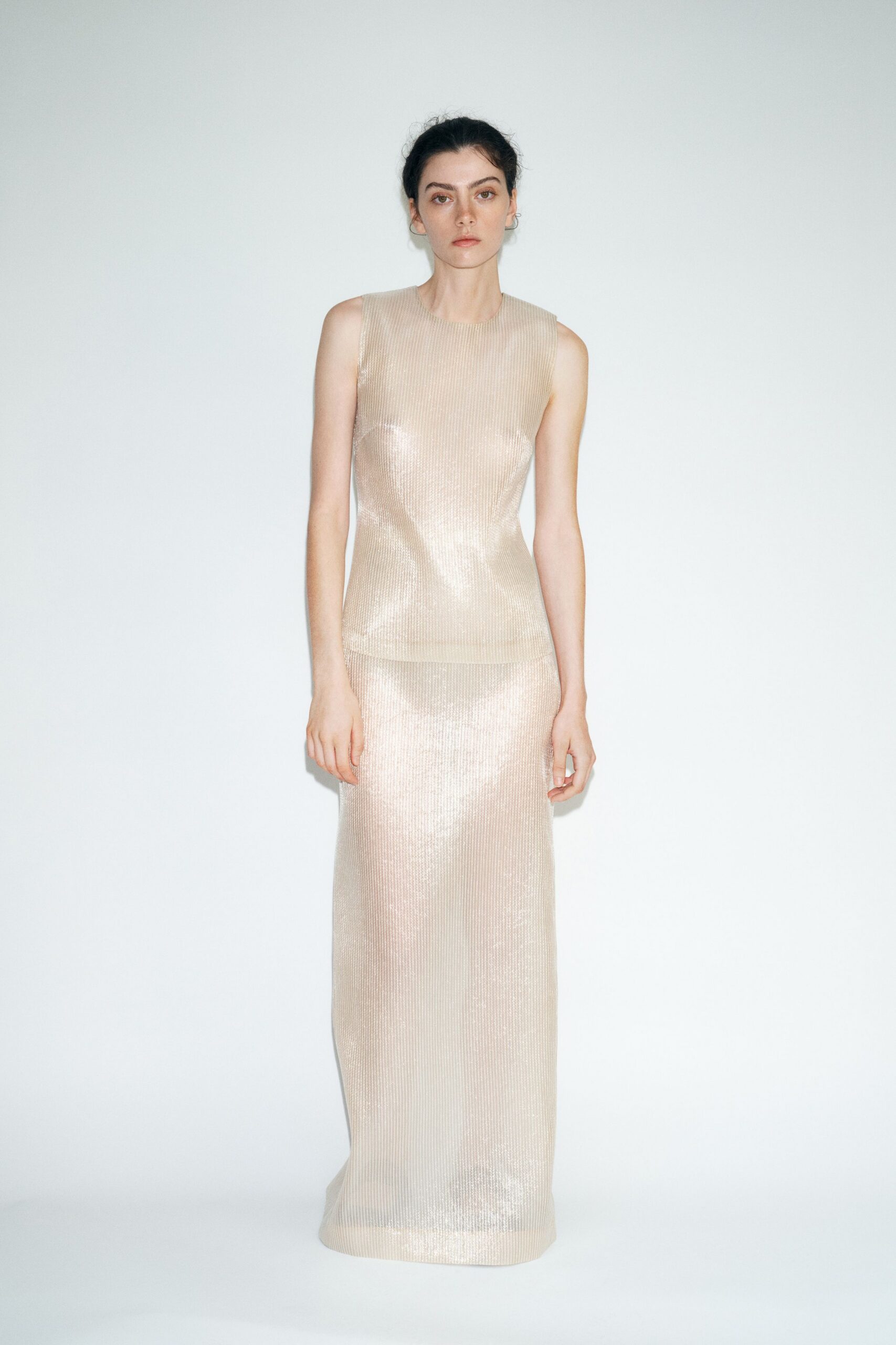 Champagne lamé shell top, fine vertical ribs catching light across the bust. Matching ankle-length column skirt in the same ribbed lamé, hem barely clearing the floor. Museum atrium at noon, every footstep bouncing off pale stone.