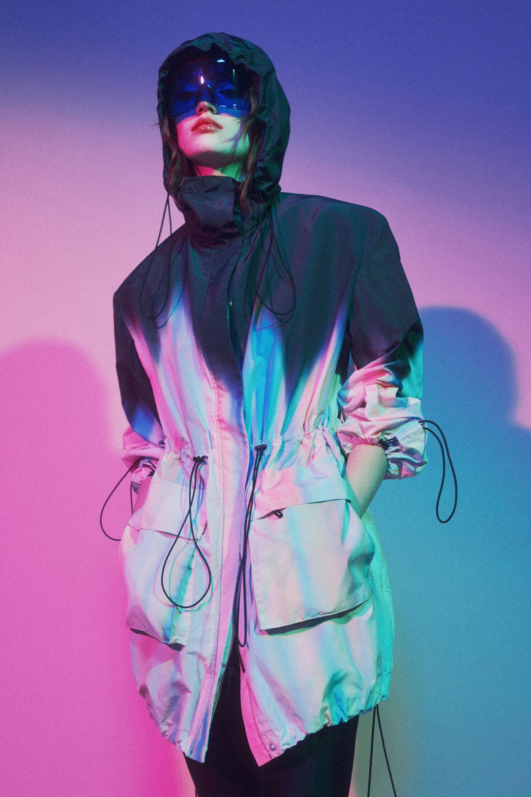 Blue mirrored wraparound visor, glossy and opaque across the eyes. Color-block nylon parka with drawcord hood, cinched waist and flap pockets. Black slim trousers, mostly hidden beneath the long hem. Waiting on a ferry deck after dark, spray needling the rail.