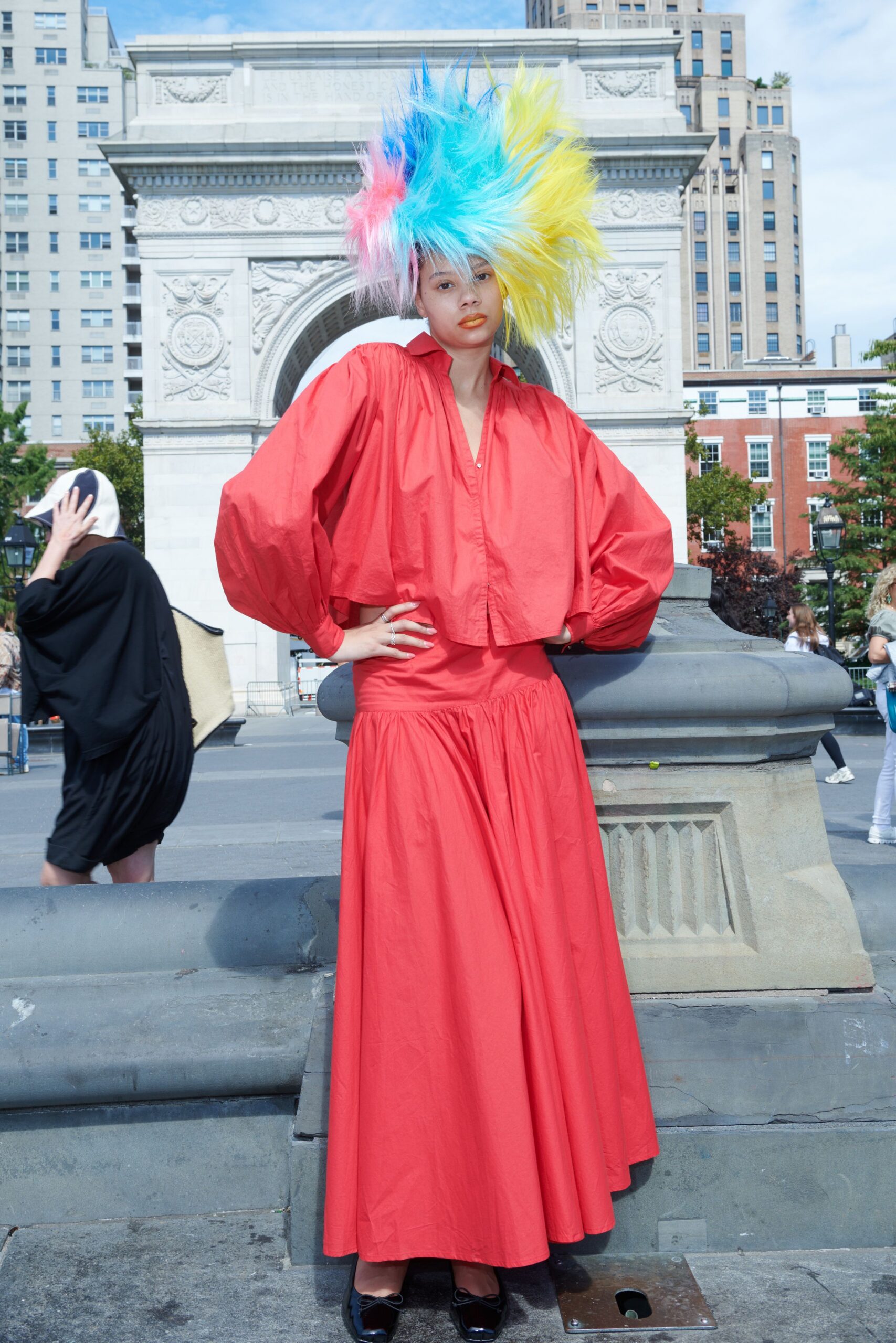 Spiked faux-fur wig in aqua, lemon and candy pink. Tomato cotton blouse with balloon sleeves and a narrow hidden placket. Matching gathered skirt, dropped waist seam softening into a full sweep. Black patent flats with tiny bow knots. Sun on the arch, buskers tuning up across the square.