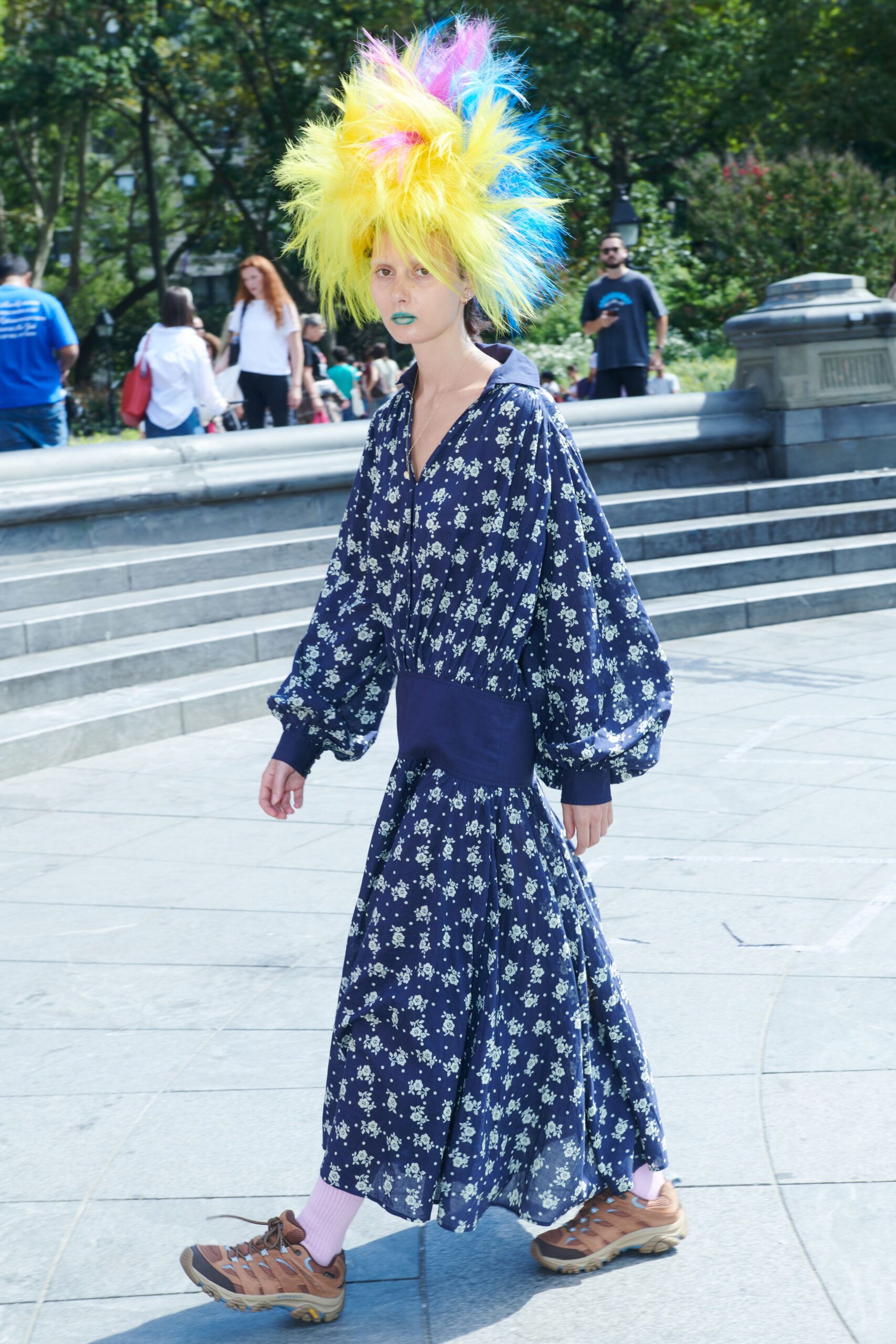 Towering neon faux-fur wig, electric blue and pink spiking through acid yellow. Navy chiffon dress scattered with tiny white florals, bloused sleeves into deep cuffs. Pale lilac ribbed crew socks. Brown mesh hikers with looped laces and thick beige tread. Crossing the park plaza after a children's party took a strange turn.