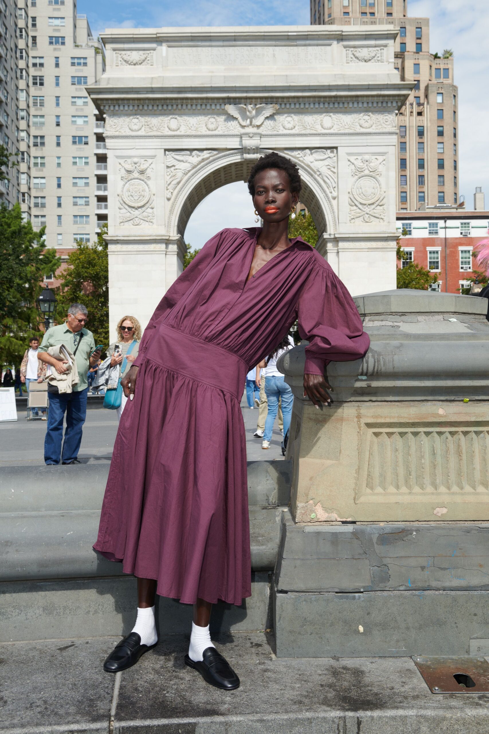 Mulberry cotton dress with knife pleats falling into a dropped gathered skirt. Small gold drop hoops brushing the jaw. White ribbed ankle socks. Black leather loafers with a blunt apron seam. Leaning by the fountain while the park chess tables fill up for noon.