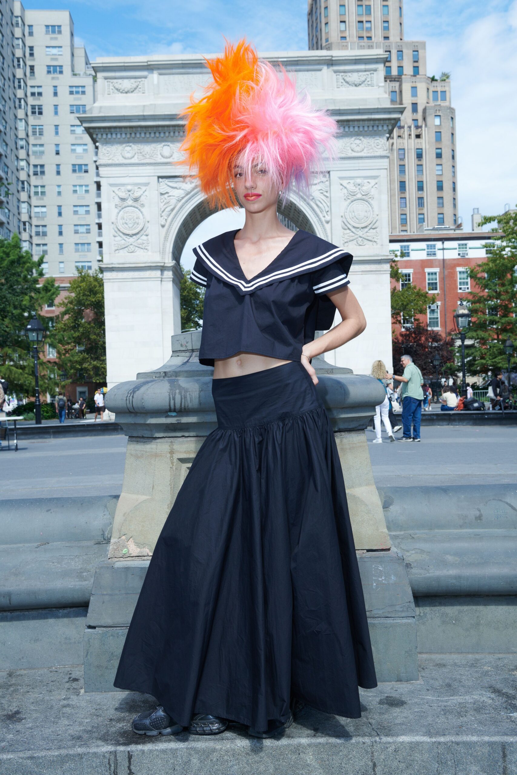 Towering shaggy wig split neon orange and bubblegum pink. Cropped navy cotton sailor top, double white stripes skimming the deep collar. Low-slung navy poplin skirt with a dropped gathered tier. Bulbous charcoal trainers peeking from the hem. Crossing the square after a costume lunch, fountain spray drying on the stone.