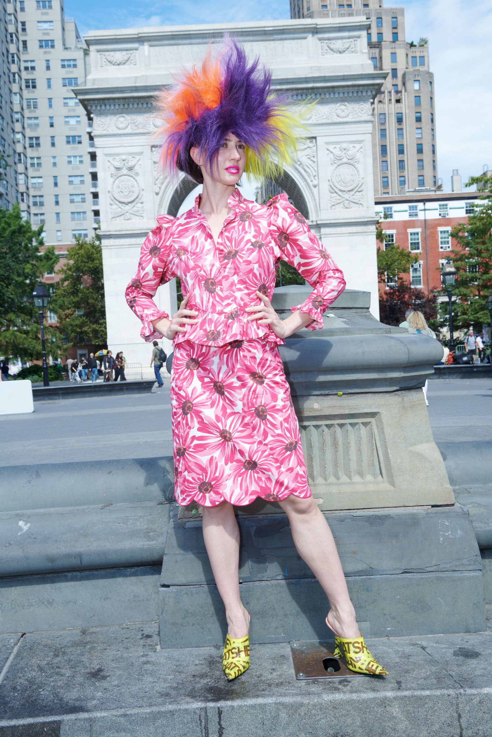 Spiked faux-fur wig in violet, tangerine and acid yellow. Pink daisy-print jacket with puffed shoulders and a peplum kick. Matching knee-length skirt, petals blown large across the satin. Chartreuse pointed mules printed all over with brown block letters. Noon in the park, heading to an outdoor chess game nobody asked for.