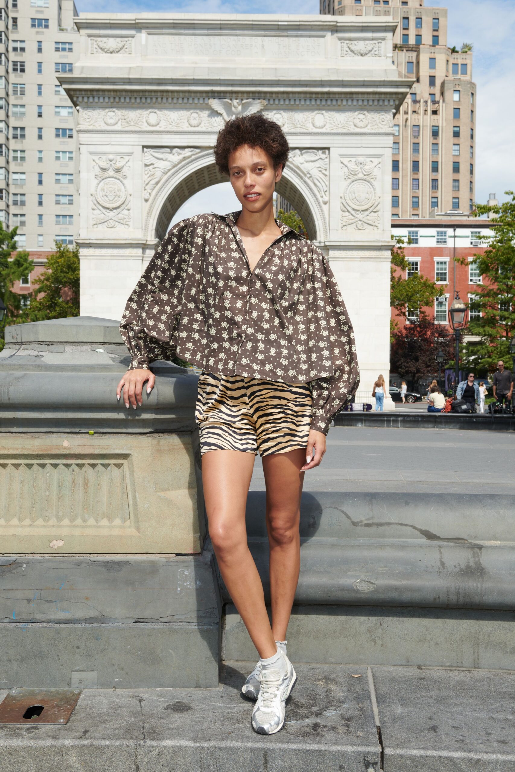 Brown floral blouse with a deep split collar and balloon sleeves. Sand zebra shorts in glossy satin, cut high on the thigh. White mesh runners with silvered toe caps. White ankle socks, softly rumpled above the shoe. Crossing the square before noon, fountain spray already lifting in the heat.
