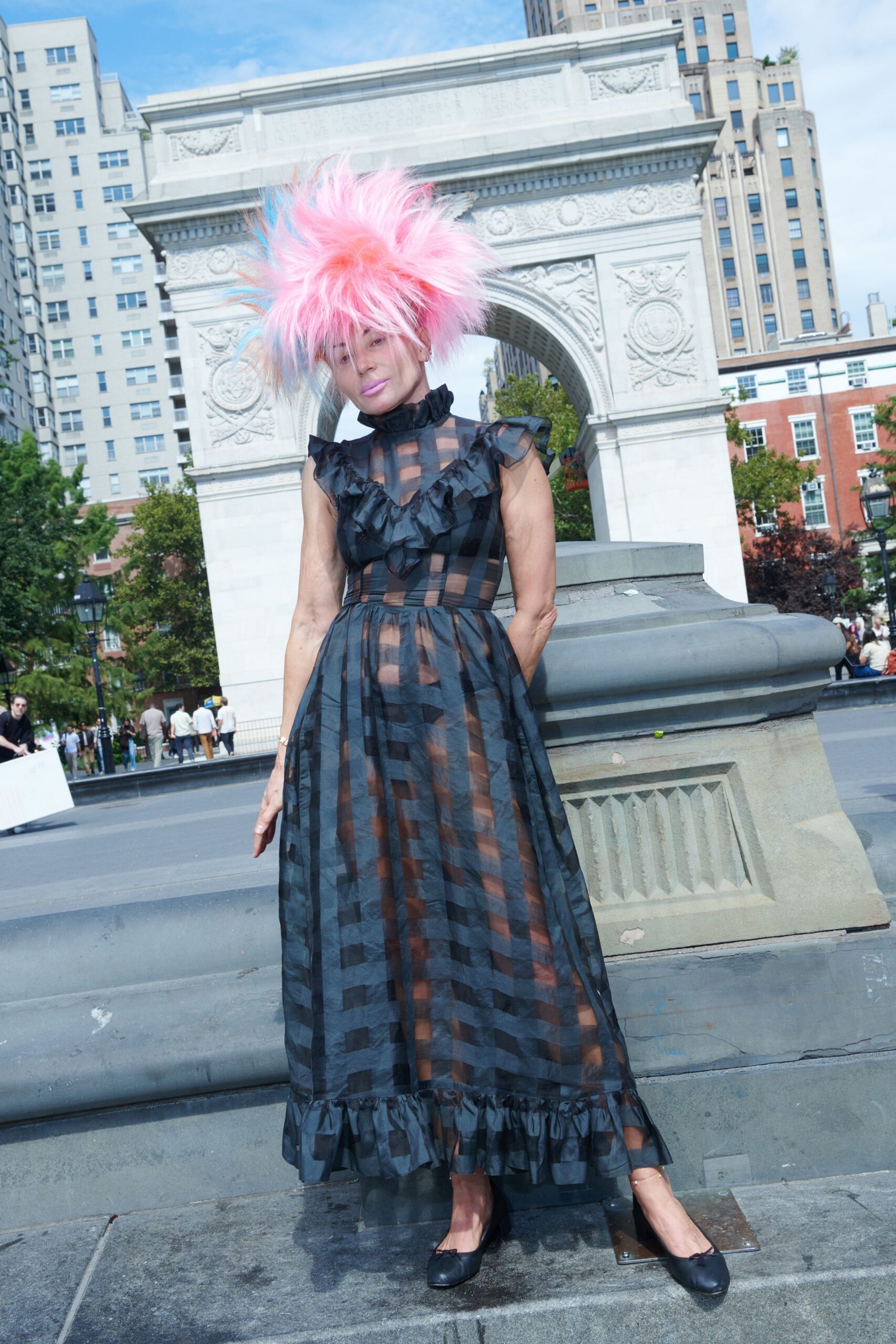 Cotton-candy pink wig blasted into blue-tipped spikes. Sheer black checked organza dress with ruffled bib and hem flounce. Black leather ballet flats with tiny bow knots. Crossing the square after noon, fountain spray catching the skirt.