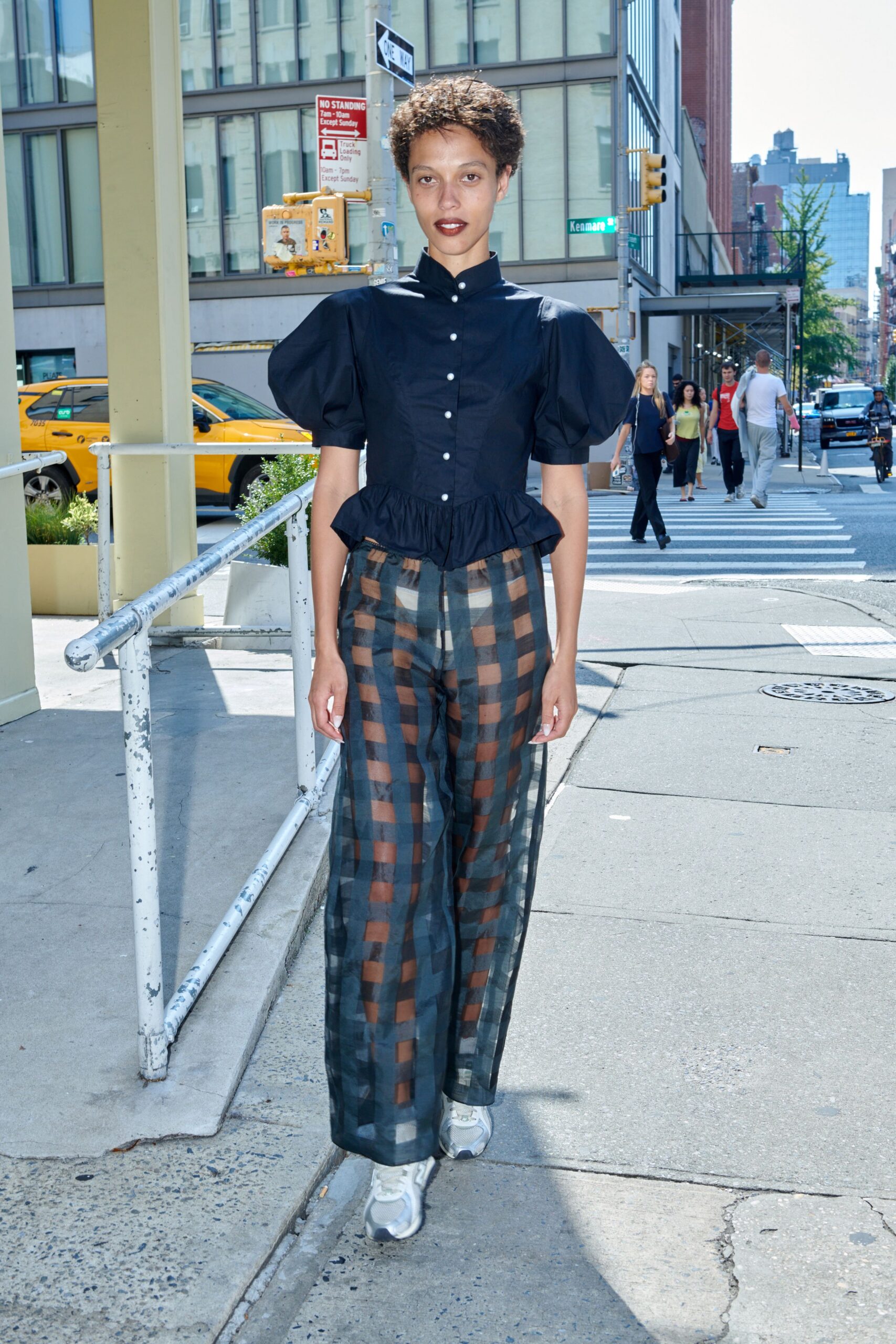 Black cotton blouse with balloon sleeves, pearl buttons, and a peplum hem. Sheer wide-leg trousers in smoky brown and slate check. Silver running sneakers with thick molded soles. Crossing for an iced coffee before the walk signal changes.