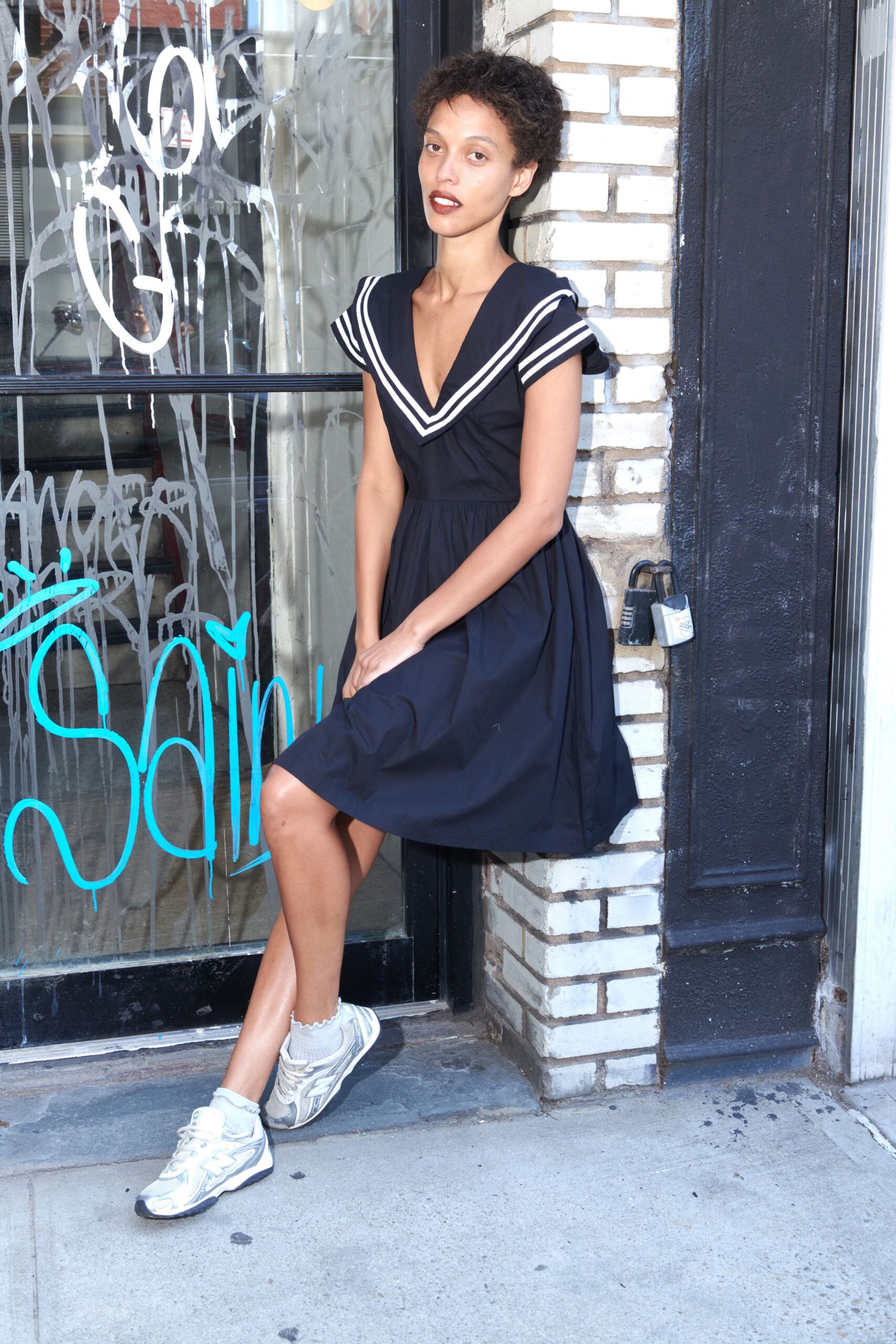 Navy cotton dress with a plunging sailor collar edged in double white stripes. White ankle socks with a lettuce-edge ruffle. Silver mesh running sneakers with thick white laces. Leaning outside the laundromat before the delicates cycle finishes.