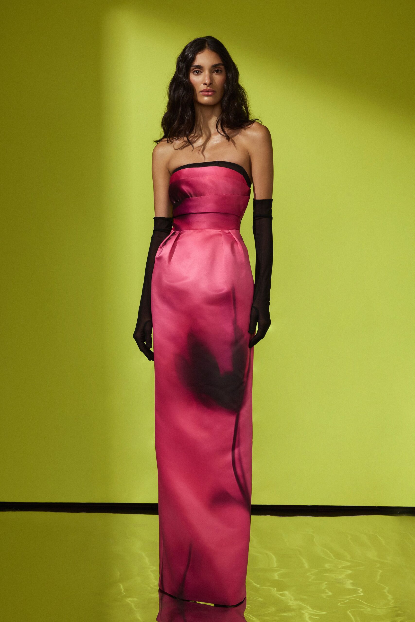 Strapless fuchsia satin column gown, black binding at the neckline and smoky shadow print. Sheer black opera gloves, seams tracing straight up the forearm. Hotel ballroom after midnight, floral centerpiece water staining the linen.