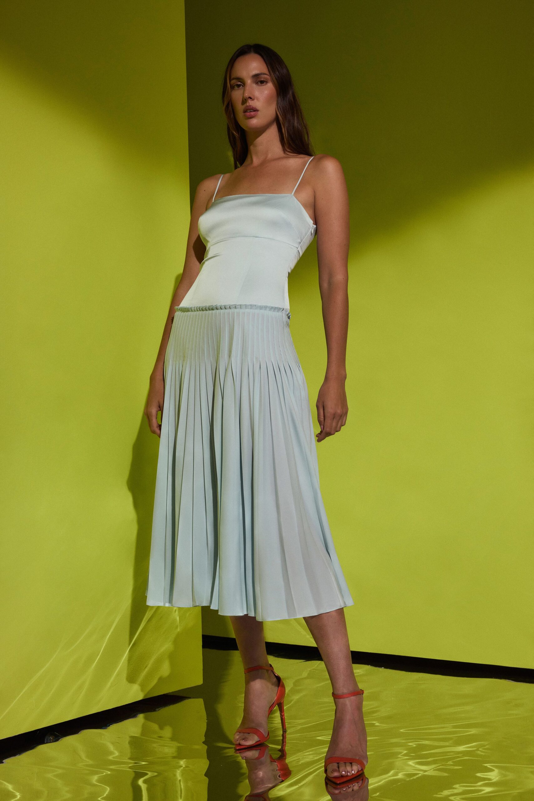 Pale celadon slip dress with a square neckline and knife-pleated drop skirt. Tangerine ankle-strap sandals with slim stiletto heels. Hotel atrium at noon, sunlight bouncing off the tiled fountain.
