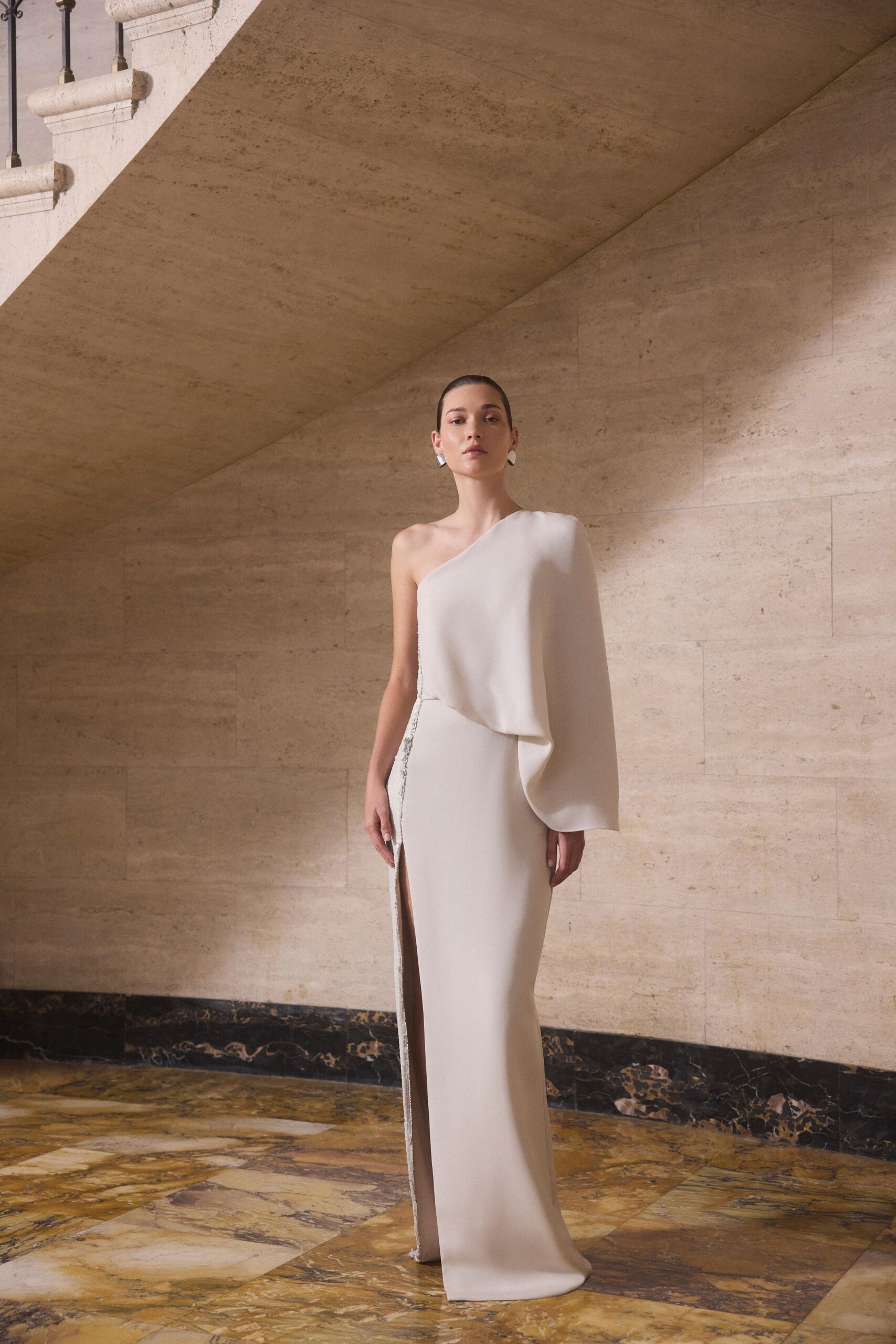 Angular silver earrings, small and bright against slicked-back hair. Chalk crepe one-shoulder column gown, one sleeve draped wide as a cape. Beaded trim glinting down the side seam into a thigh-high slit. Crossing a marble foyer before the wedding guests are called upstairs.