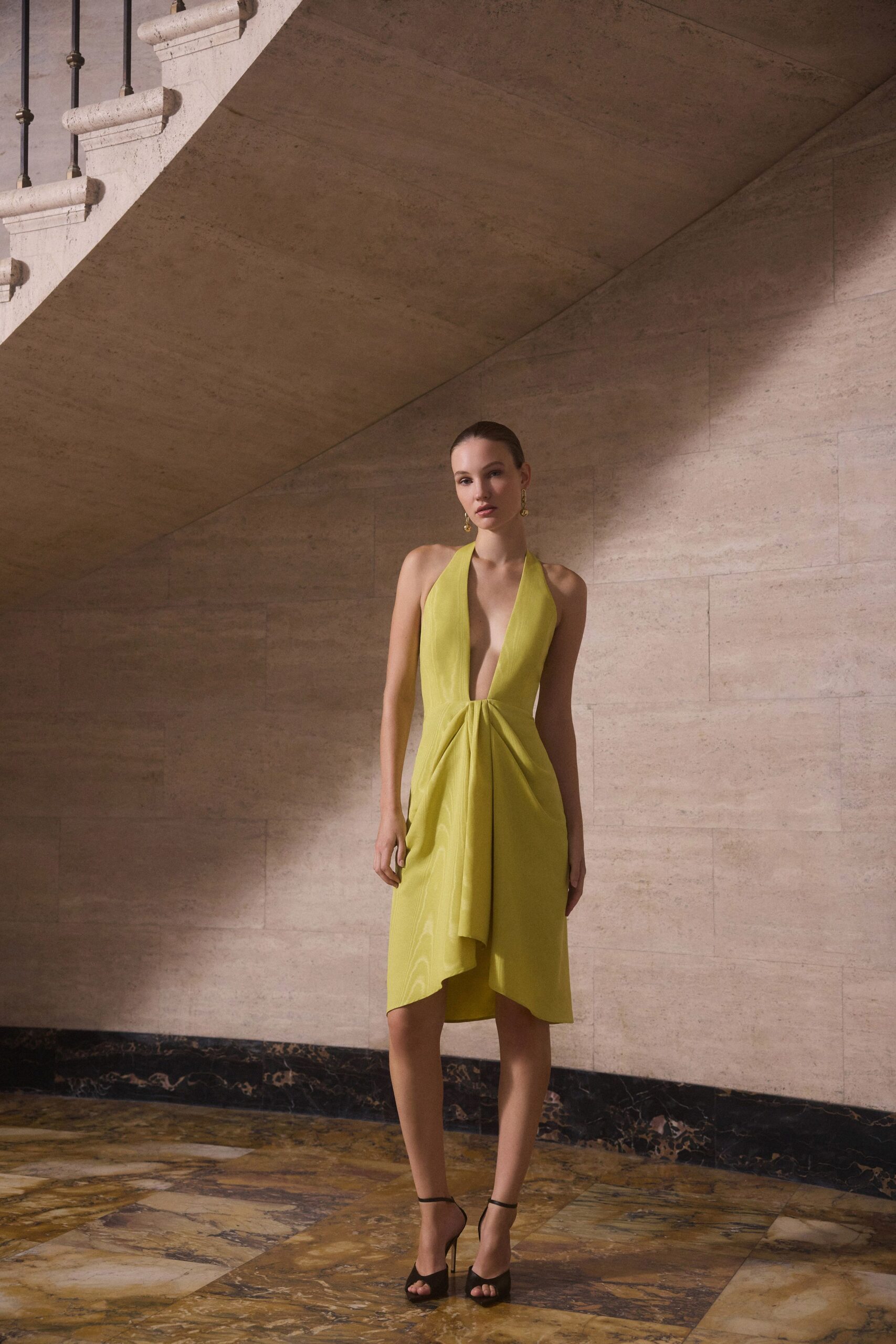 Chartreuse halter dress with a plunging V and twisted drape at the waist. Small gold drop earrings. Black ankle-strap sandals with slim stiletto heels. Crossing a hotel atrium before the wedding guests spill out for champagne.
