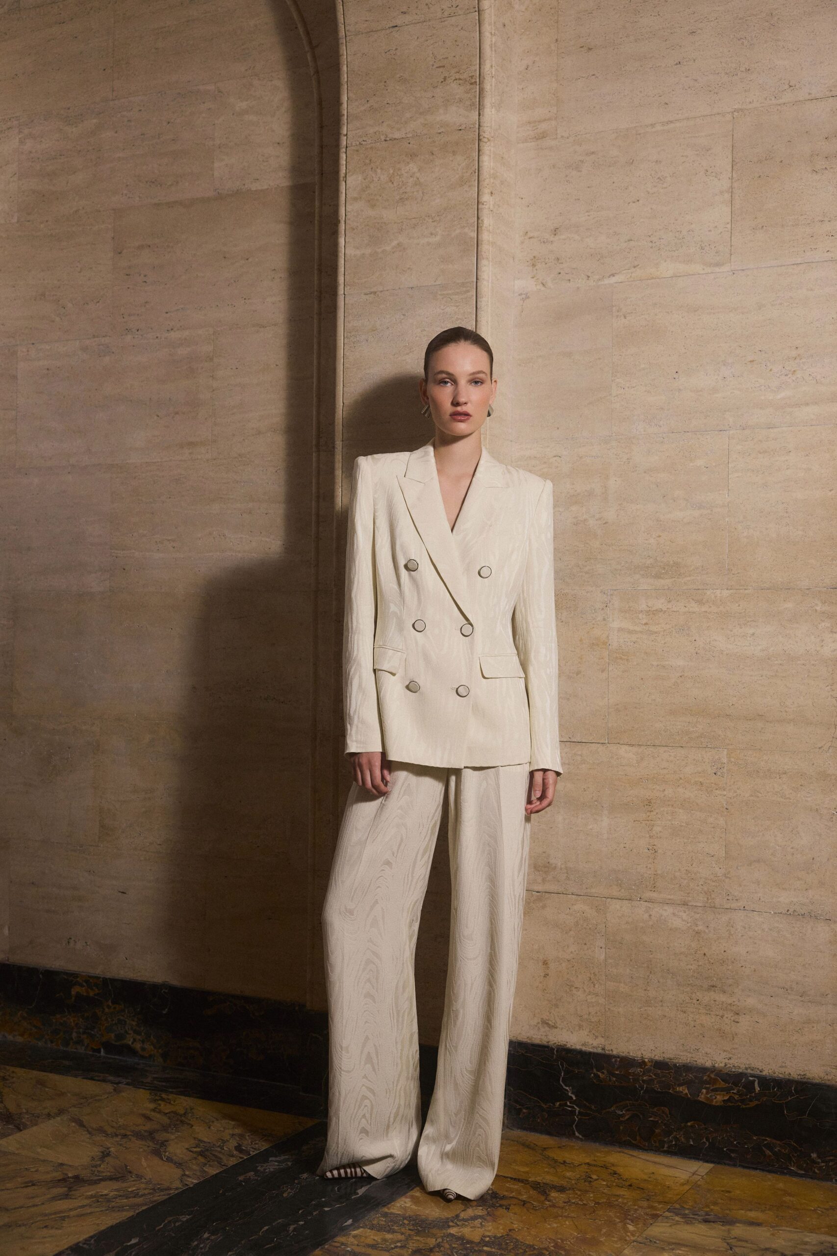 Ivory double-breasted blazer in moire, six ring-edged buttons catching the light. Matching moire trousers falling long enough to pool over the shoes. Striped pointed heels peeking from the hem. Waiting out a wedding in a stone foyer after the champagne ran dry.