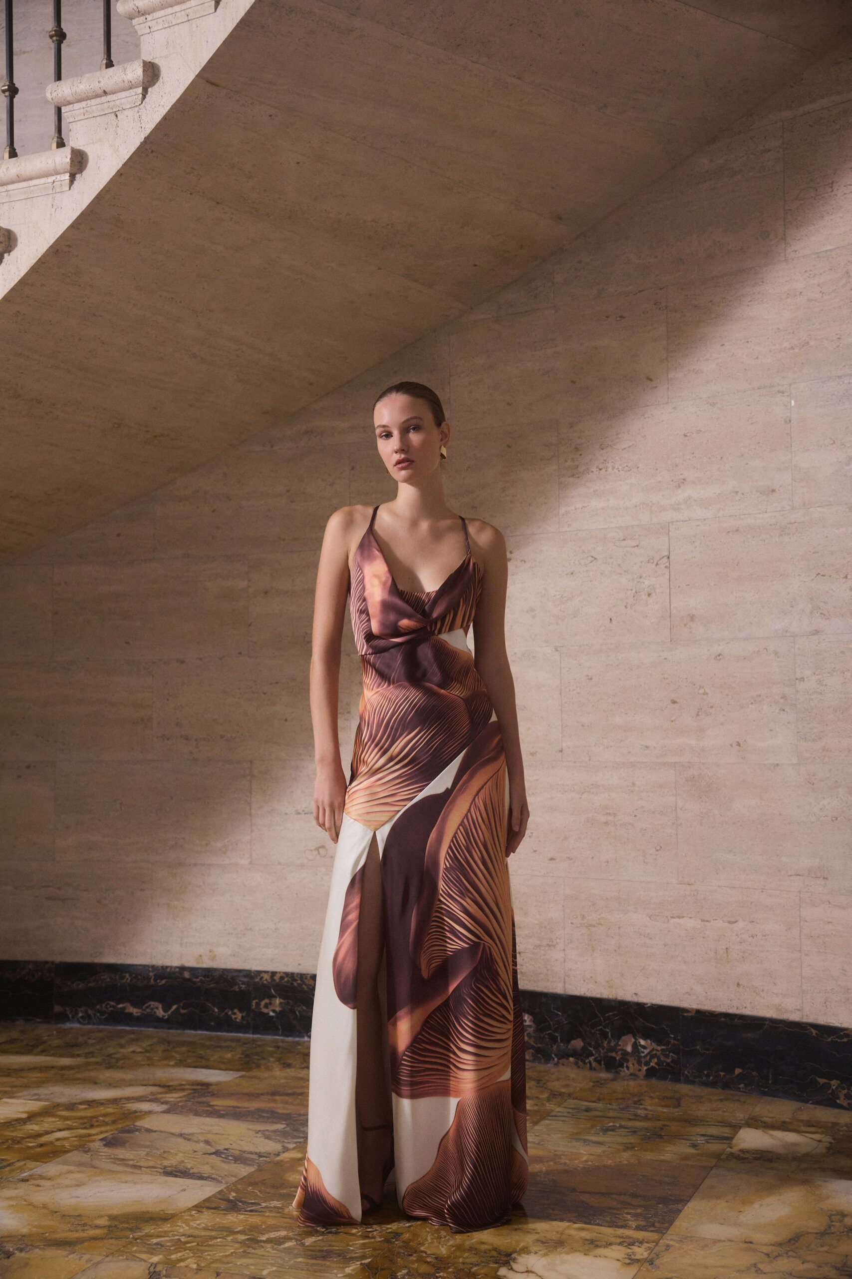 Bias-cut satin slip gown in chestnut and cream, draped cowl bodice and thigh slit. Small gold geometric drops. Dark barely-there straps. Sunlit museum stairwell, arriving early enough to hear your own heels.