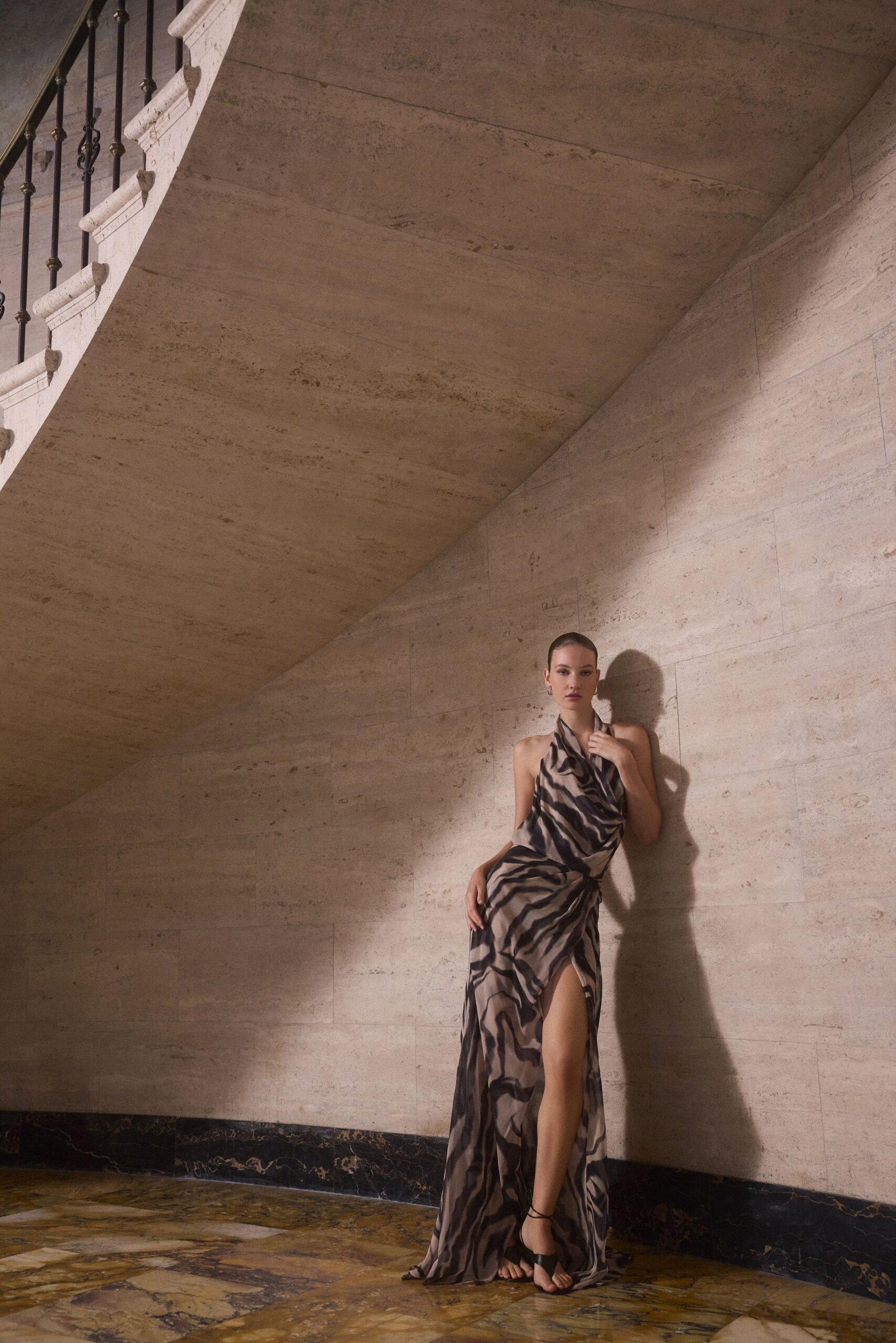 Smoke-grey draped halter gown in zebra-striped silk, thigh slit knotted at the hip. Black thong sandals with thin wraparound ankle ties. Small silver hoops. Crossing a marble lobby after midnight, still warm from the terrace.