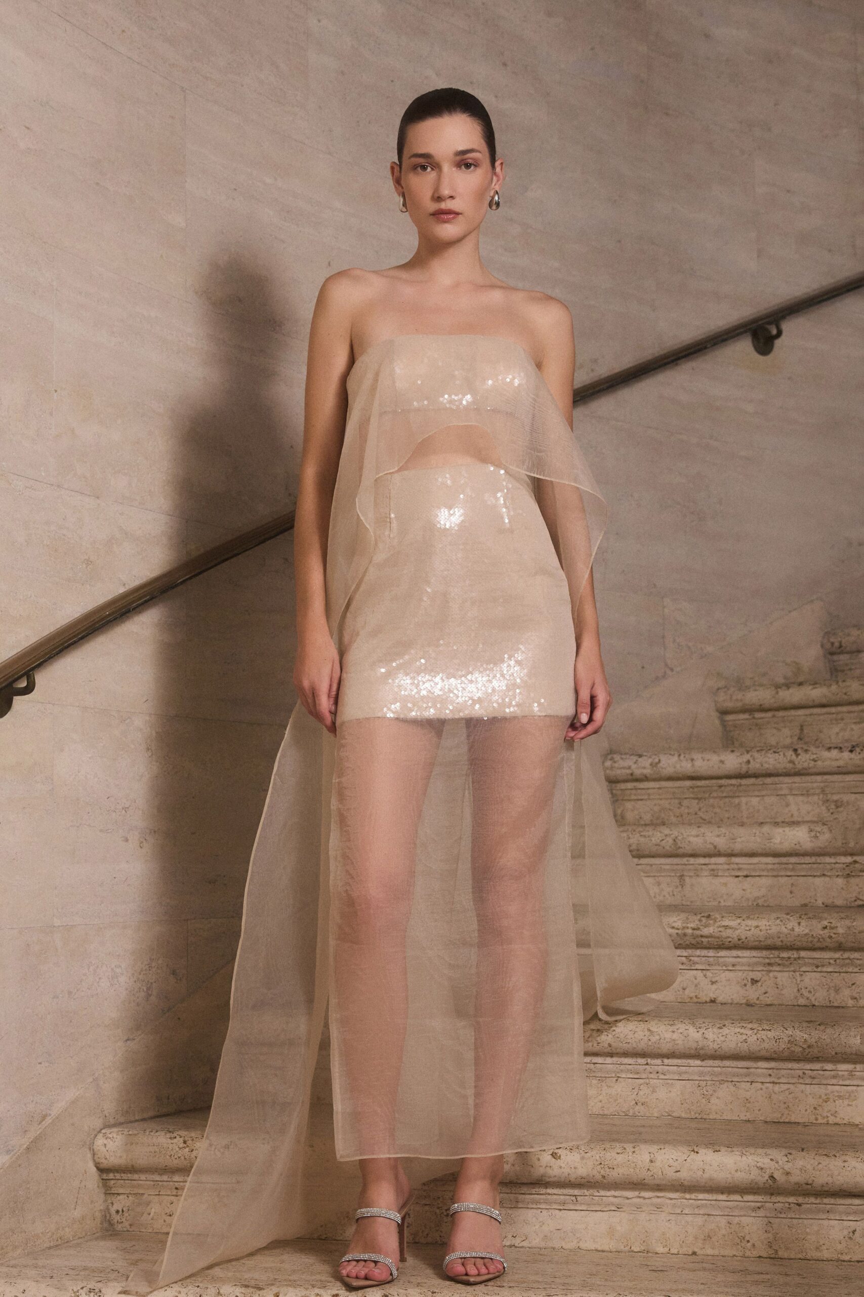 Champagne sequin column mini under a sheer organza panel falling to the floor. Chunky silver hoops. Crystal twin-strap sandals with slim stiletto heels. At the top of a museum staircase after everyone else has gone downstairs.