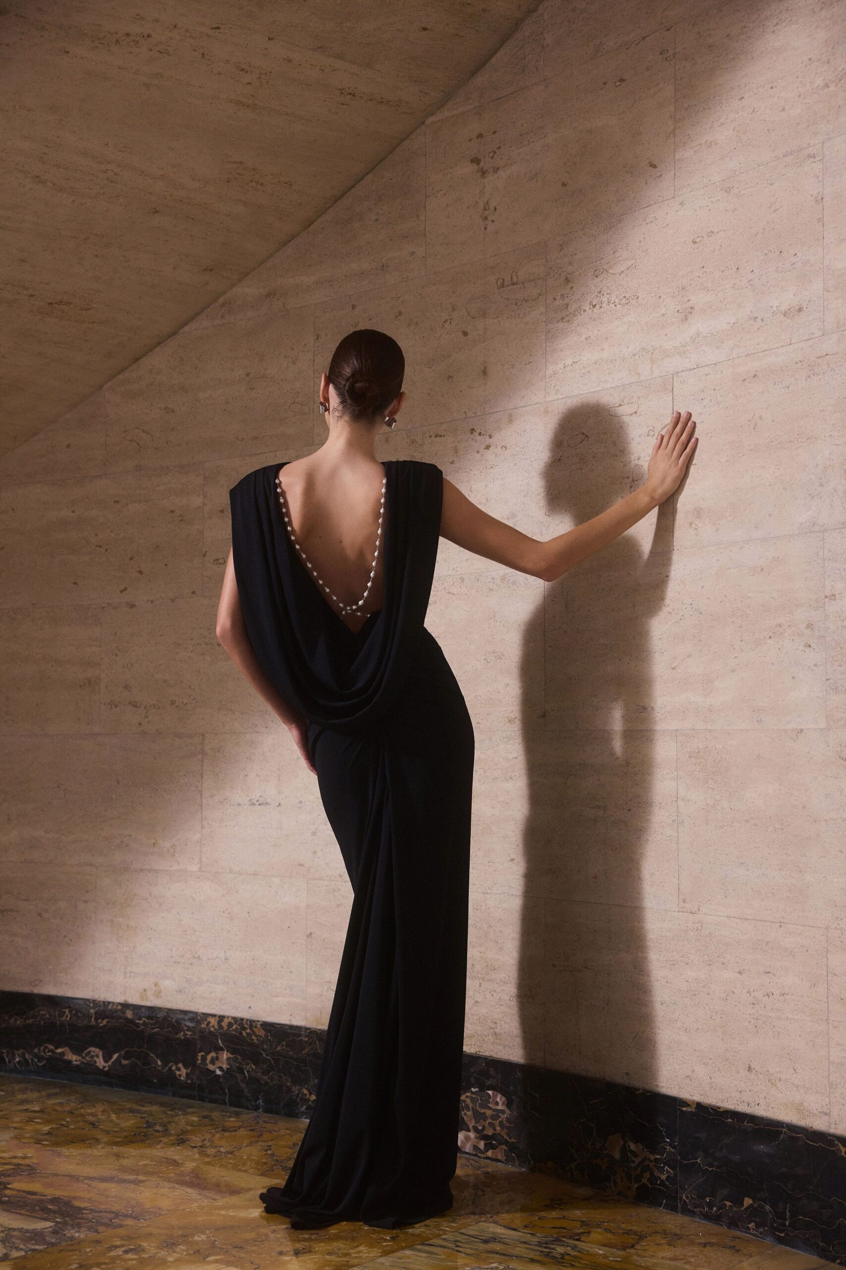 Black column gown in liquid jersey, back plunged to a pearl-trimmed cowl. Small silver block earrings. Pausing on a marble stair landing after everyone else has gone in.