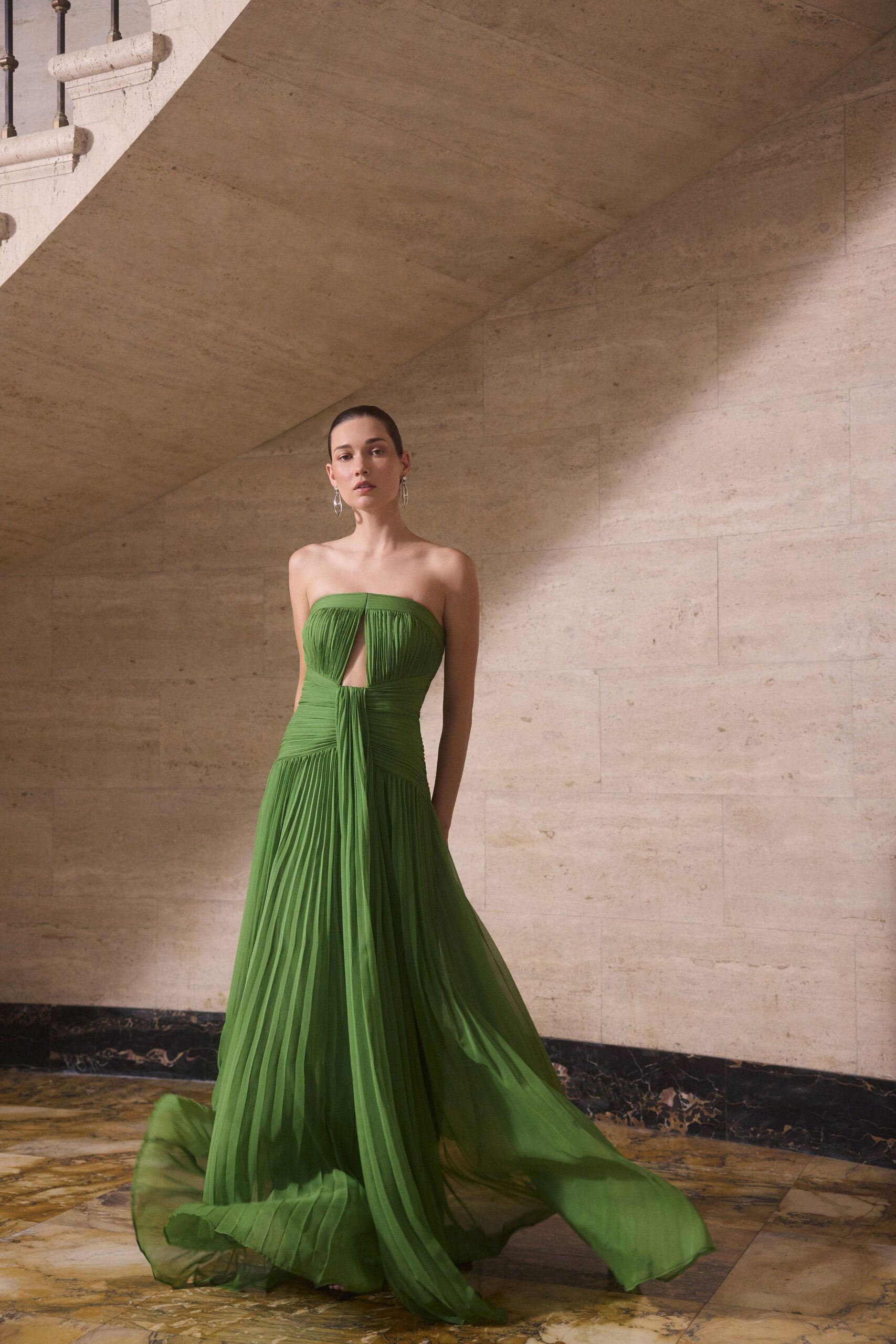 Strapless leaf-green chiffon gown, sunray pleats falling from a ruched waist and narrow front cutout. Long silver drop earrings. Crossing a museum atrium just after the doors have closed.