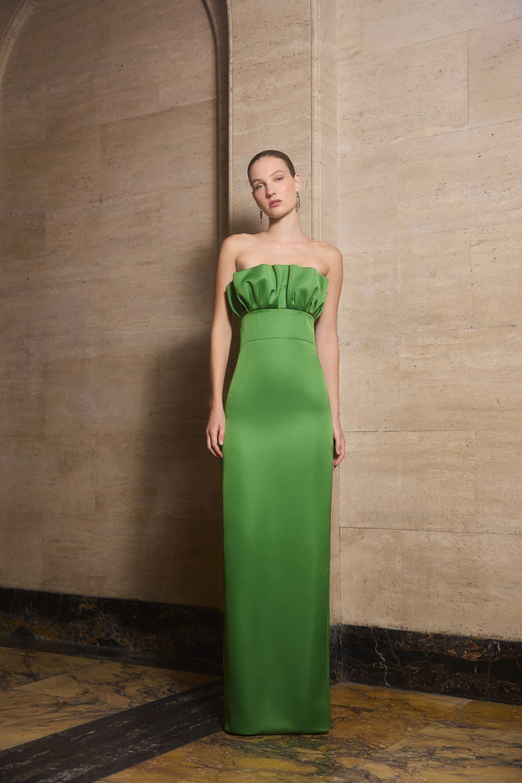 Strapless parakeet-green column gown with a puffed taffeta bust and wide waistband. Long silver drops brushing the jaw. Under a limestone arcade before the wedding guests are let upstairs.