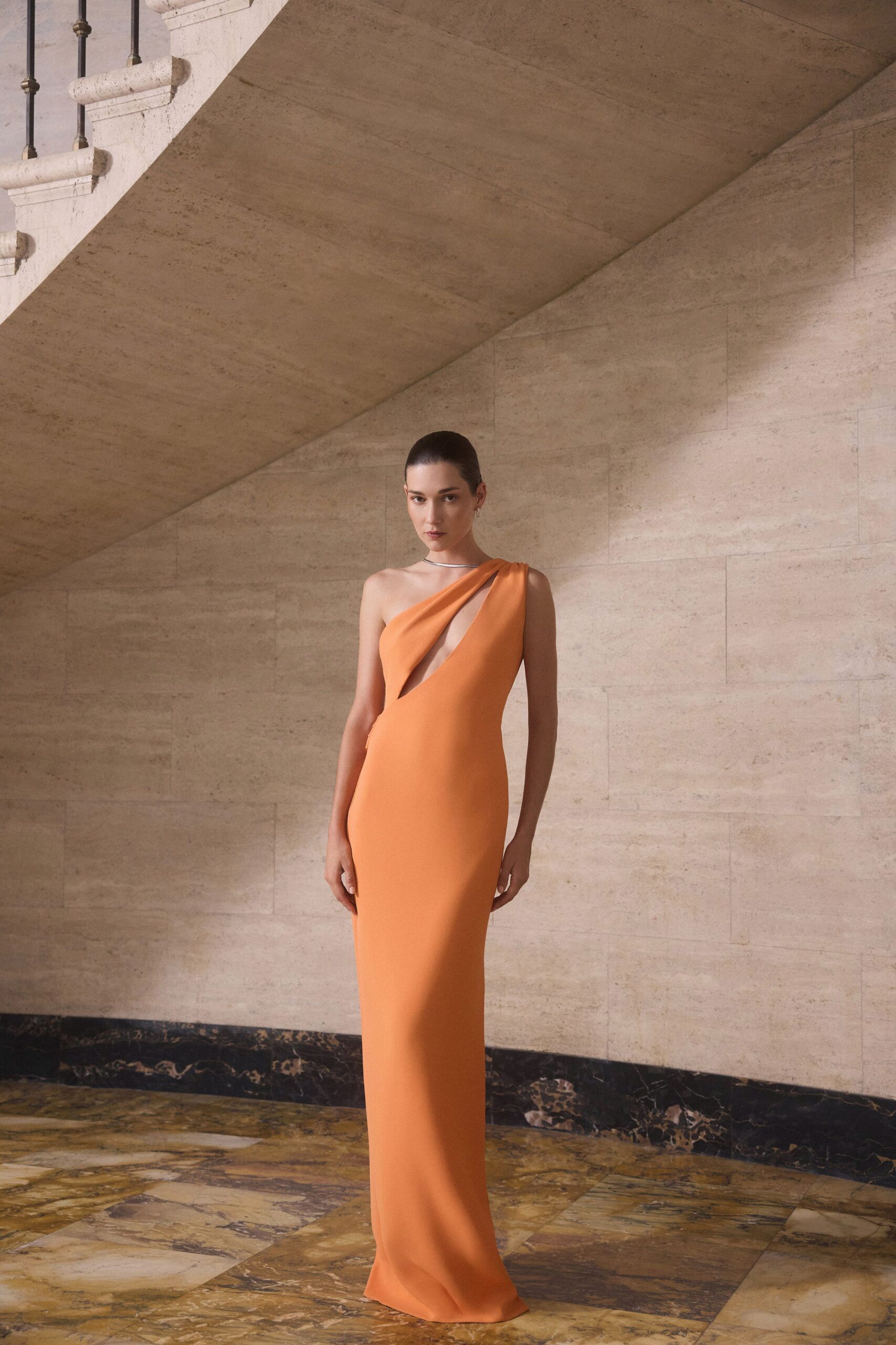 Apricot column gown with one-shoulder drape and a sharp teardrop cutout. Slim silver collar sitting close against the throat. Tiny drop earrings. Crossing a stone hotel lobby before the wedding guests come down.
