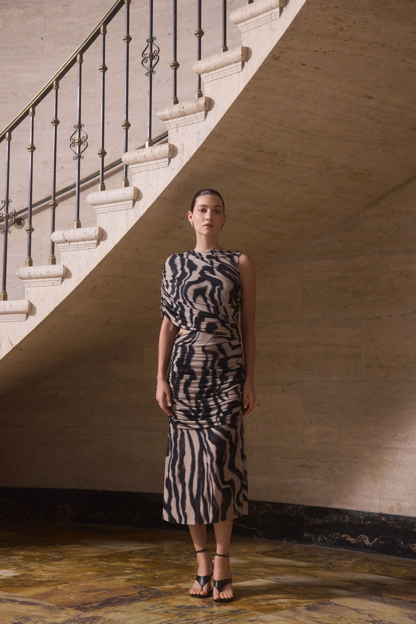 One-shoulder ruched jersey dress in blurred zebra stripes, side cutout at the waist. Small silver hoop drops. Black leather toe-loop sandals with fine ankle straps. Crossing a marble foyer before the wedding guests drift upstairs.