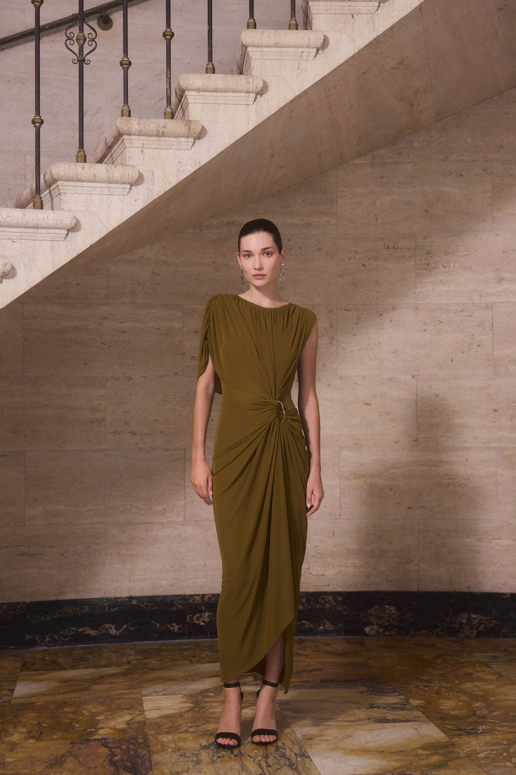 Olive jersey column dress, gathered from neckline into a ring-twisted waist. Dangling silver loops. Black ankle-strap sandals. Crossing a marble lobby after the ceremony, heels ticking under the stairs.