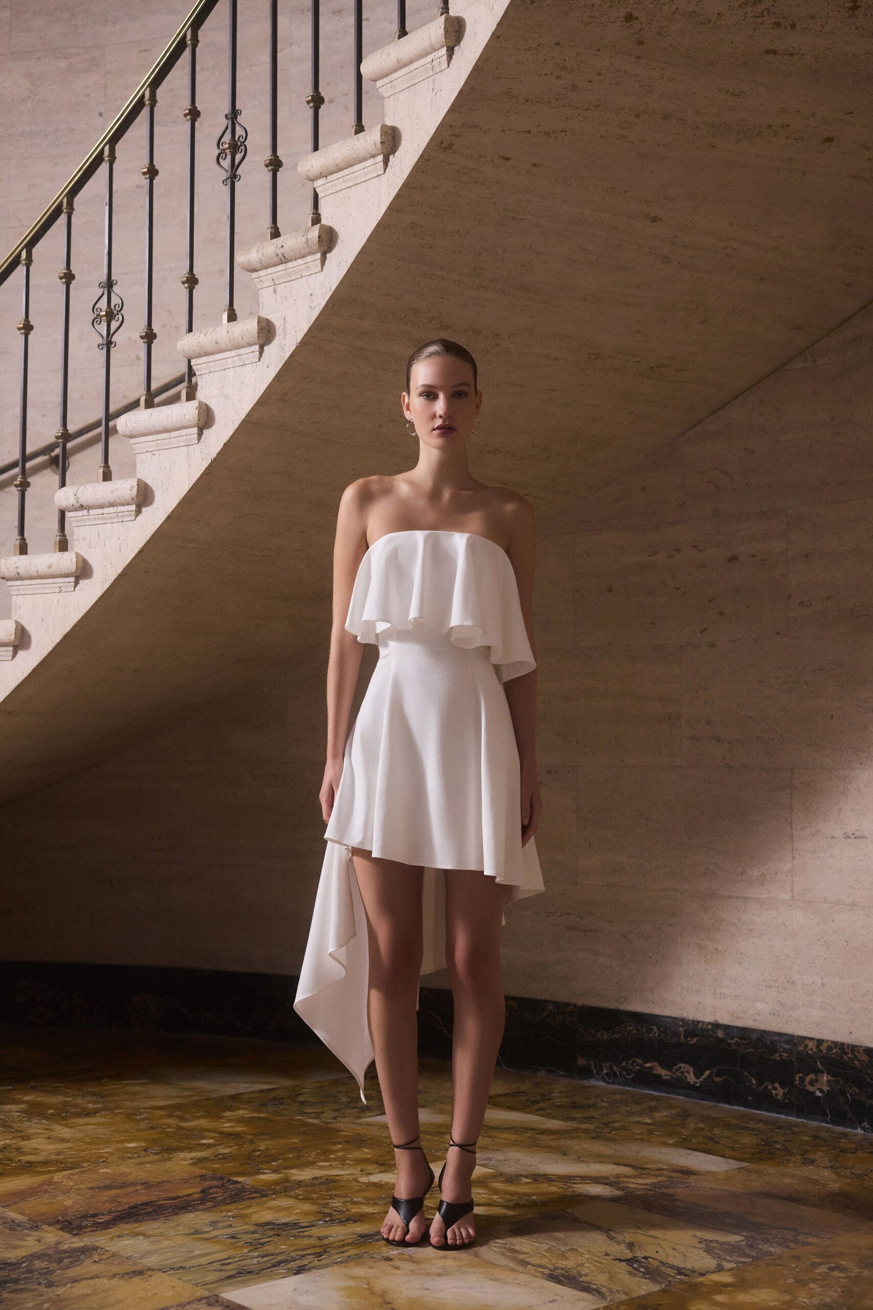 Strapless white dress with a floating flounce and one long back drape. Black leather ankle-strap heels with a narrow toe band. Small silver hoop drops. At the bottom of a stone stair after the ceremony has emptied out.