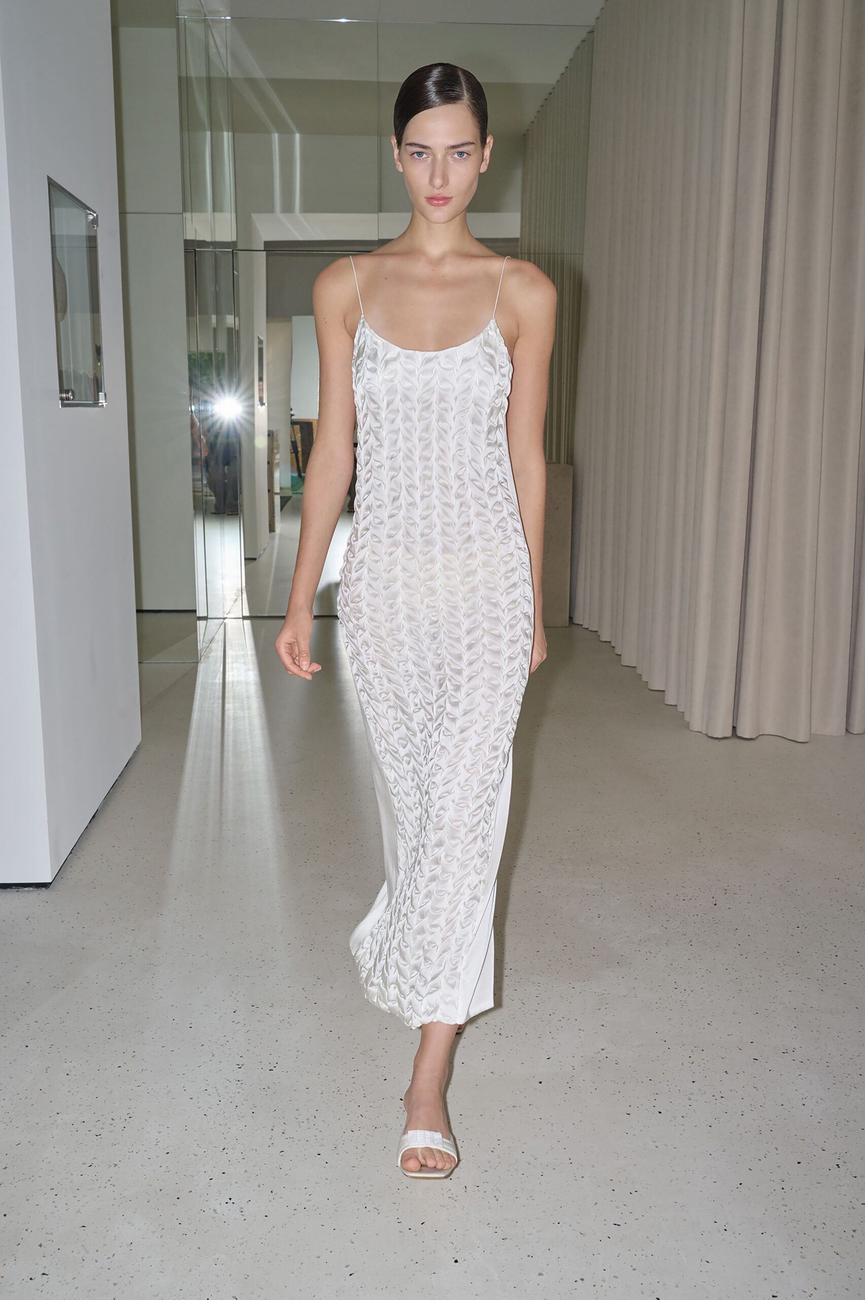 Ivory column slip dress with ruched petal-texture panels and fine straps. White square-toe slides with a softly padded band. Sunlit hotel corridor, heading downstairs before anyone else wakes.