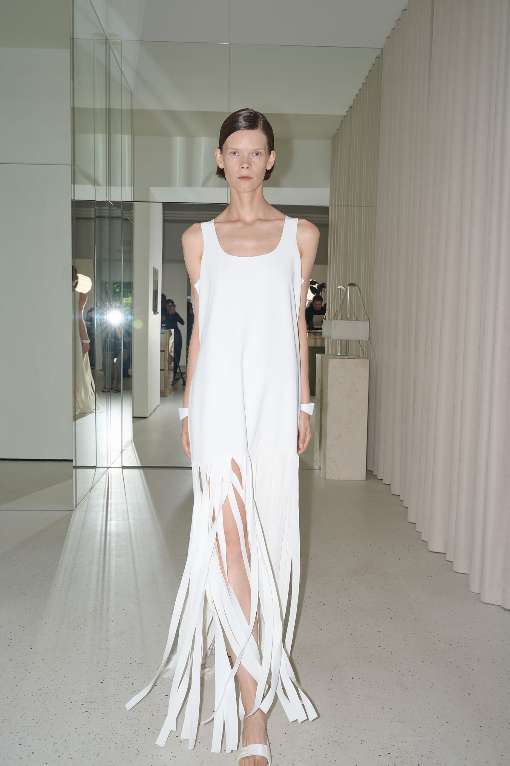White crepe column dress with a scooped tank neckline and sliced fringe hem. White cuff bracelets, blunt and paper-clean at each wrist. White slide sandals with a single padded strap. Crossing a silent atrium just after the cleaners finished the marble.