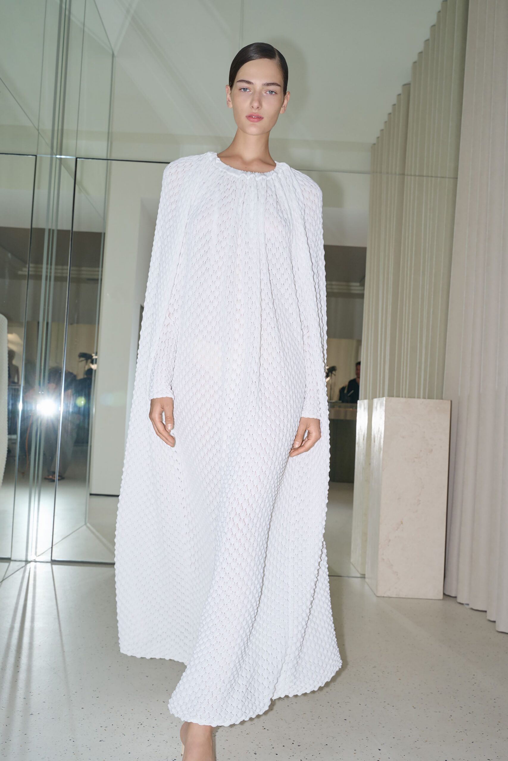 Floor-length white gown in puckered honeycomb knit, gathered softly at the neck. Crossing a marble lobby before anyone has started speaking.