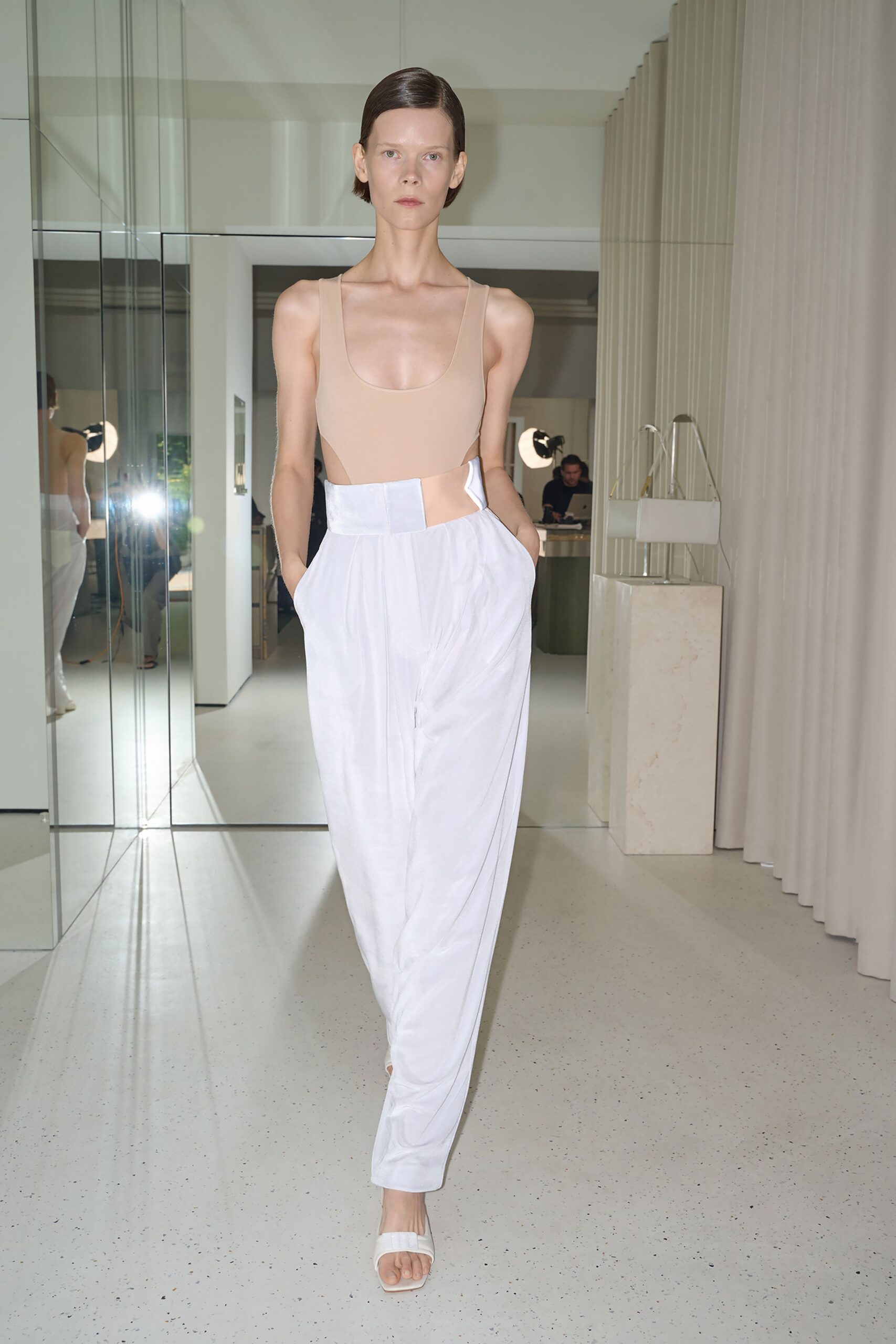Nude stretch bodysuit with a deep scoop neck and open side cutaways. White fluid trousers with a broad satin waistband and soft front drape. White slide sandals with a square toe. Walking through a silent showroom before the first appointment arrives.