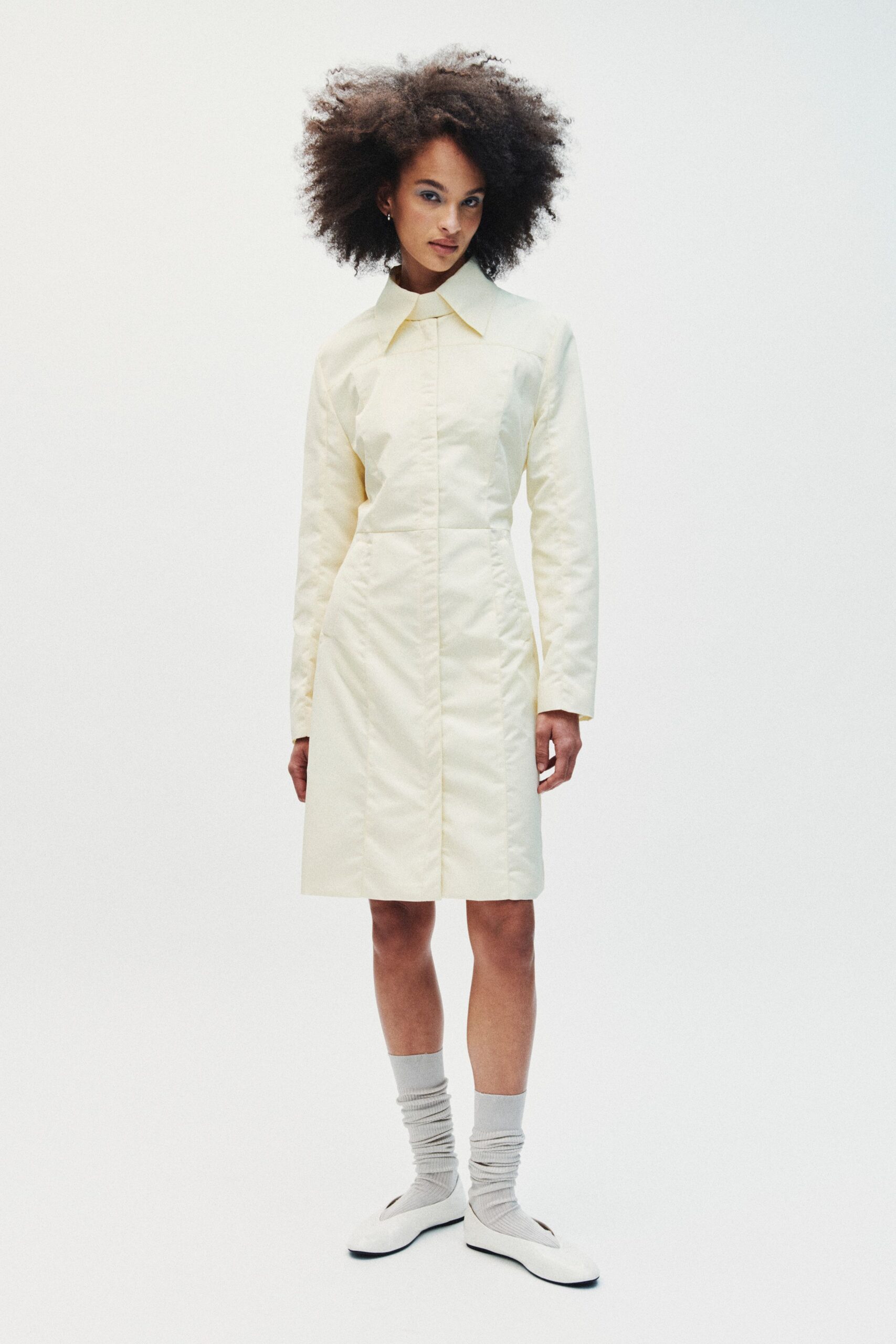 Pale butter shift dress in crisp satin, pointed collar and center seam. Light grey ribbed socks, softly slouched above the ankle. White wedge flats with squared toes and black outsole peeking. School hallway before first bell, floors just waxed.
