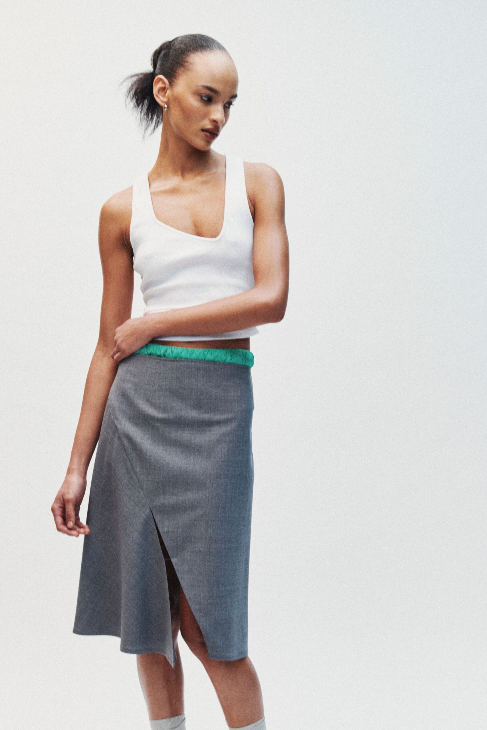 White ribbed knit tank with a deep square scoop neckline. Grey suiting midi skirt, wrapped askew with a sharp front slit. Bright jade elastic peeking above the skirt. Pale grey knee socks. Office tower atrium, lunch break spent circling the dry fountain.
