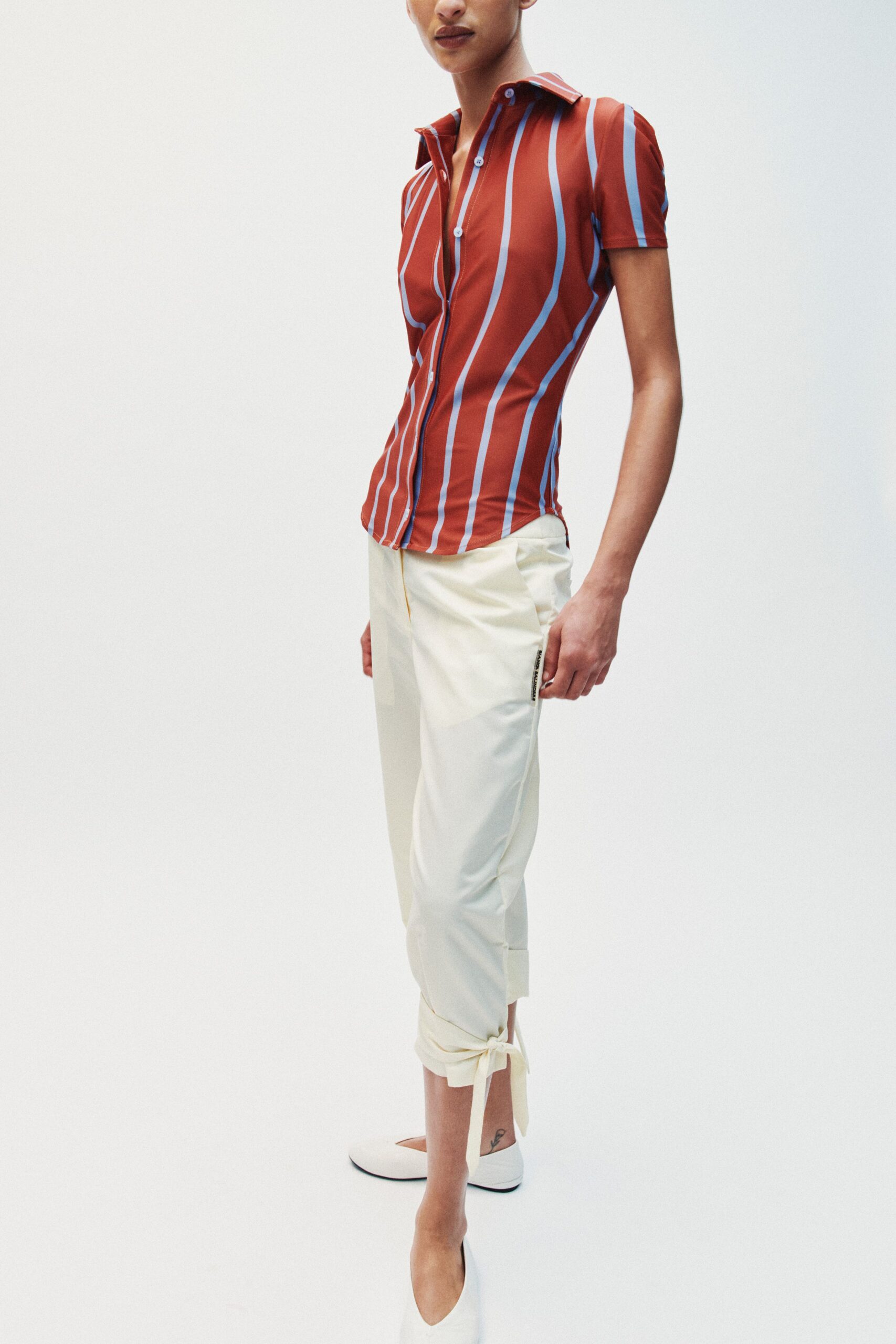 Rust stretch shirt with pale blue warped stripes and a long pointed collar. Cream cropped trousers, cuffs rolled and tied shut above the ankles. White pointed flats with a clean vamp. Walking out for hotel breakfast after sleeping in yesterday's makeup.