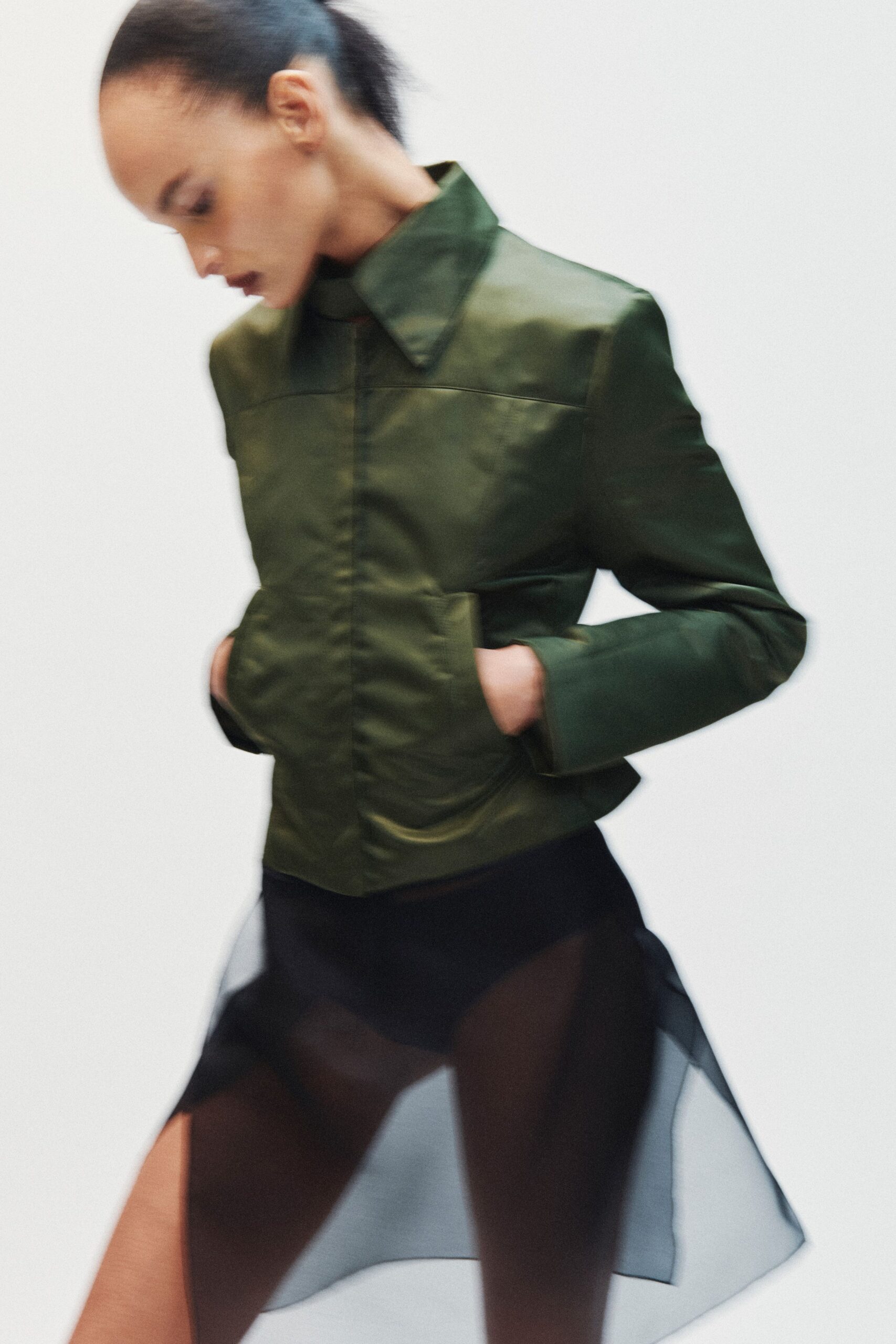 Bottle-green satin jacket, concealed placket and pointed collar standing high. Black sheer skirt with a brief underlayer and drifting organza panels. Dark sheer tights. Leaving a museum atrium while the glass doors fog behind you.