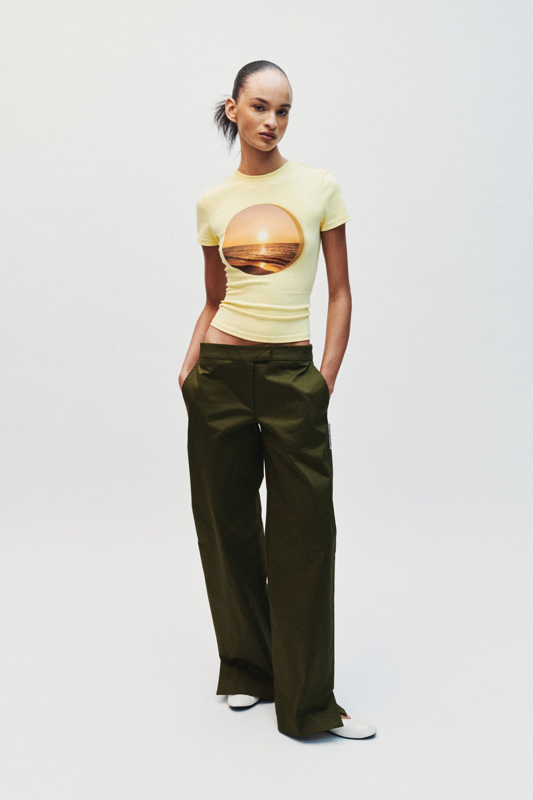 Pale lemon stretch tee with a round sunset seascape printed across the chest. Olive wide-leg trousers in crisp twill, waistband sitting low and square. White square-toe flats peeking from the pooled hem. Walking back from the beach arcade after the sky has gone violet.