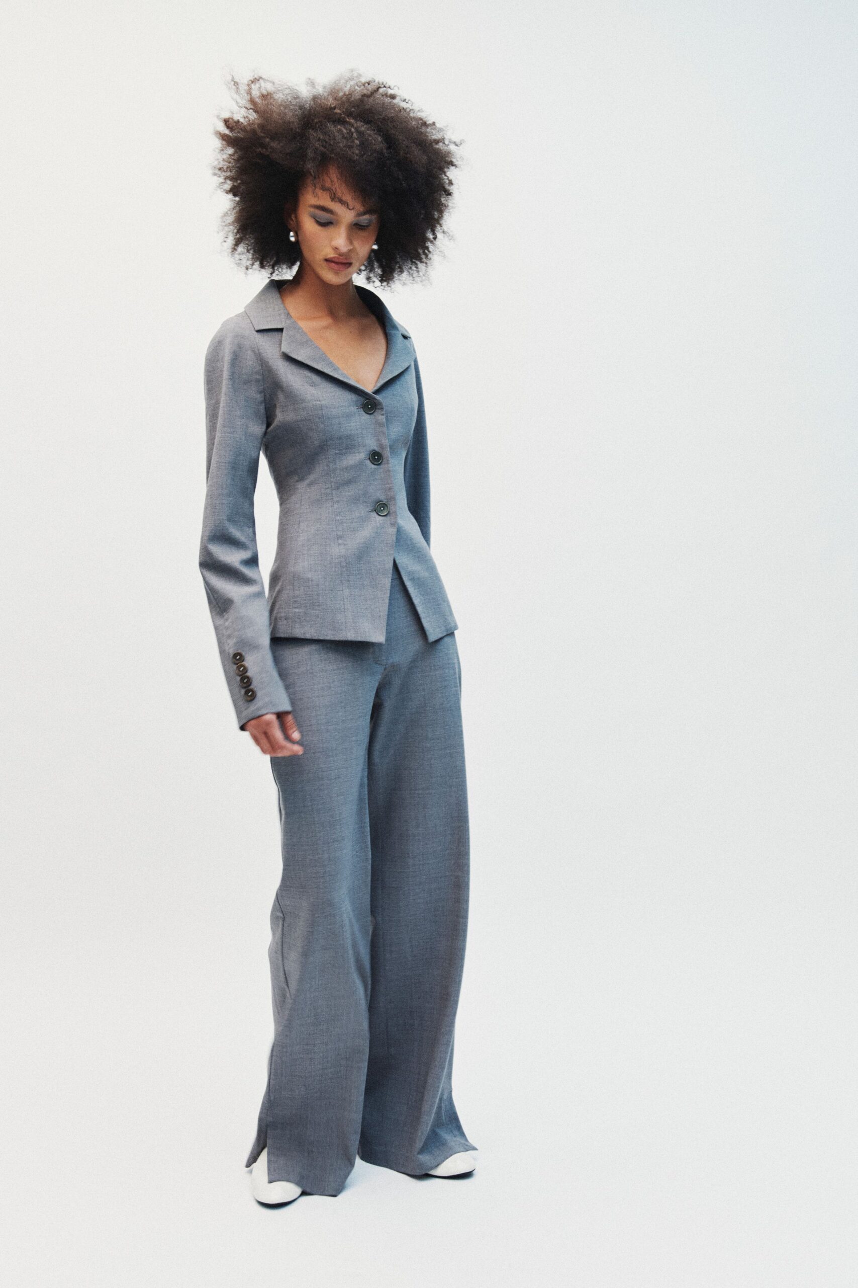 Small pearl drop earrings. Steel-grey suiting jacket, cut close with a low lapel and dark horn buttons. Matching wide-leg trousers breaking softly over the shoe, front slit at the hem. White square-toe shoes with black sole edge. For the first meeting after lunch, rain still silvering the office windows.