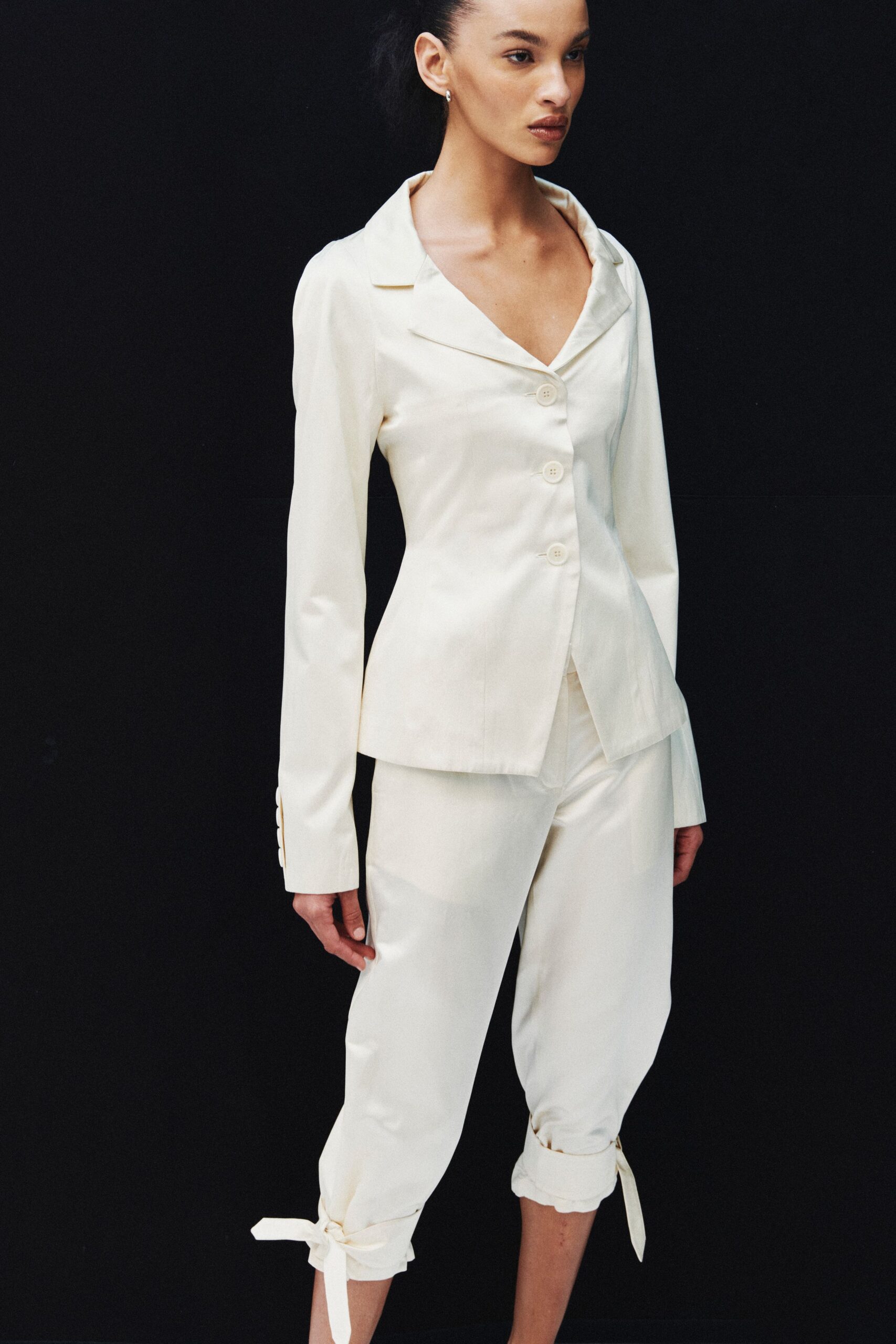 Ivory satin jacket with an off-kilter notch collar and three covered buttons. Matching satin trousers cropped at the calf, tied with long ankle bows. Small silver hoops. For signing papers at noon, then changing your mind in the stairwell.
