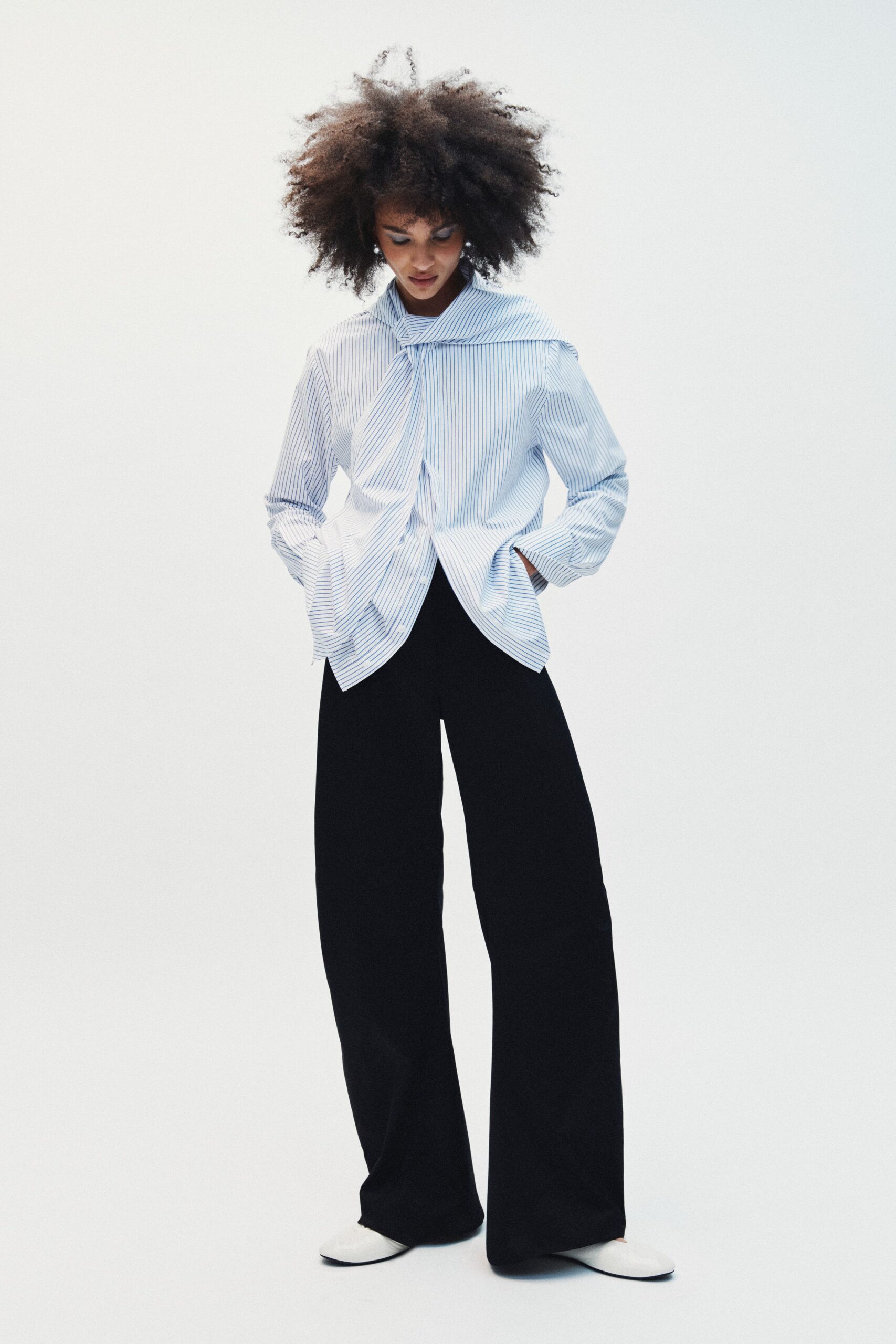 Blue-and-white banker-stripe blouse with a scarf collar knotted off-center. Black wide-leg trousers falling heavy and straight over the shoes. White pointed flats with a low vamp. First train in, coffee cooling beside the office lobby ficus.