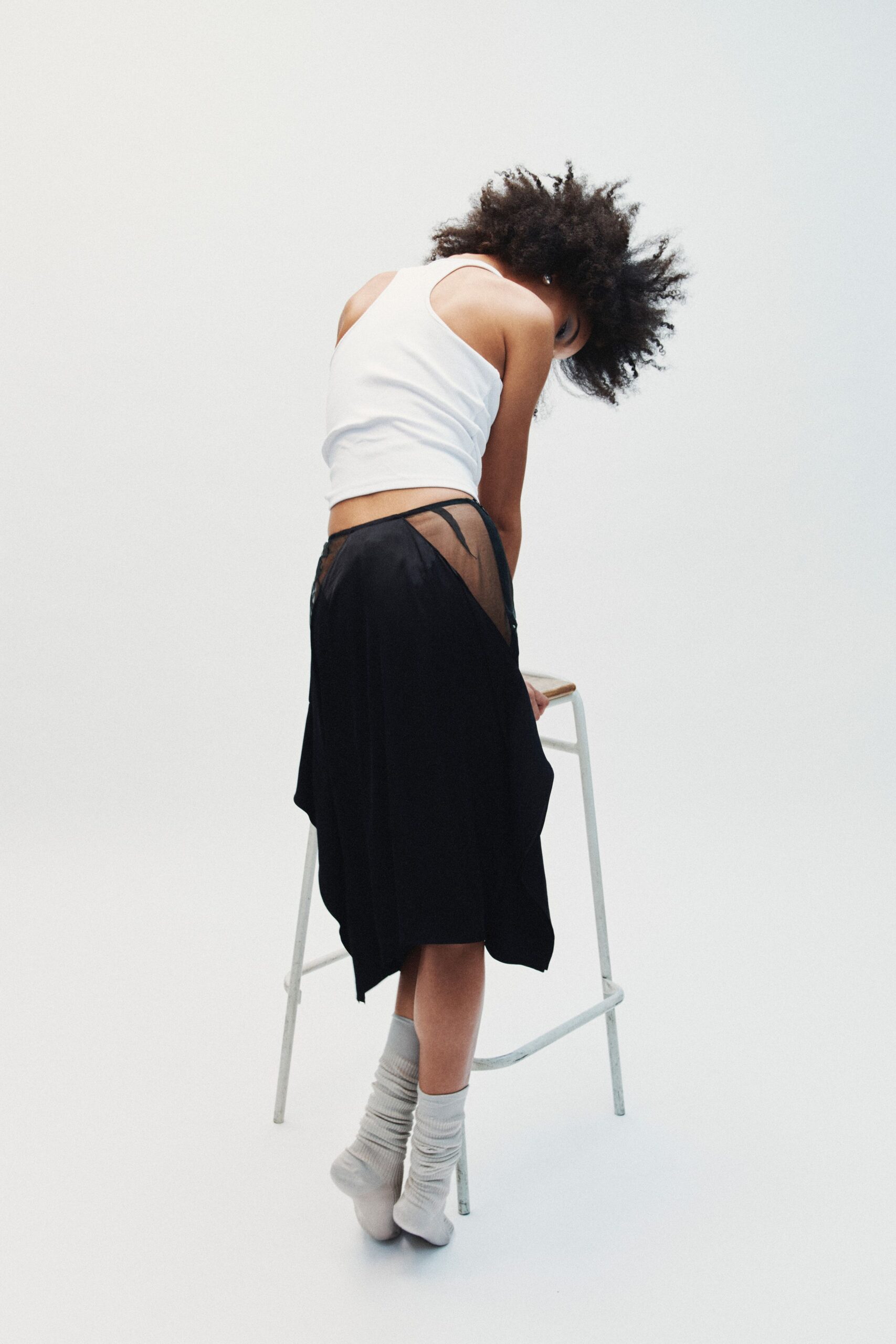 White ribbed tank, cut close with a deep racerback. Black draped skirt with sheer mesh hip panels and binding at the edge. Pale grey ribbed socks, softly pooled at the ankles. Leaning on a studio stool while the fan is still running.
