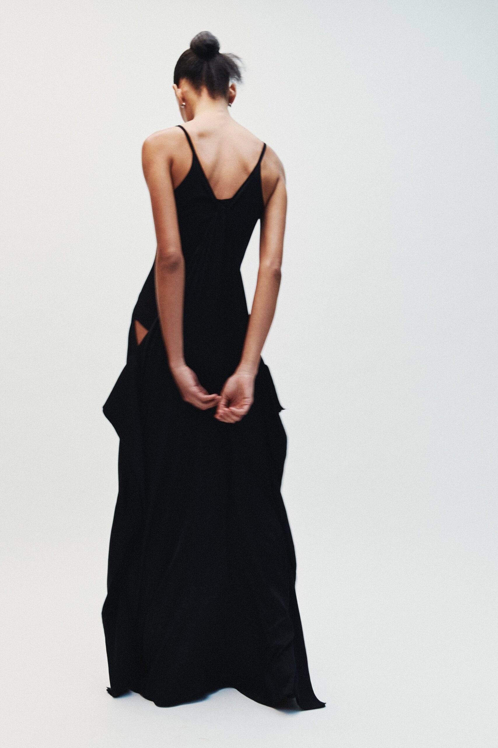 Tiny silver hoops at both ears. Black floor-length slip dress, low V back and gathered hip drape with a side cutout. Whatever you wear home after slipping out of the wedding before speeches.