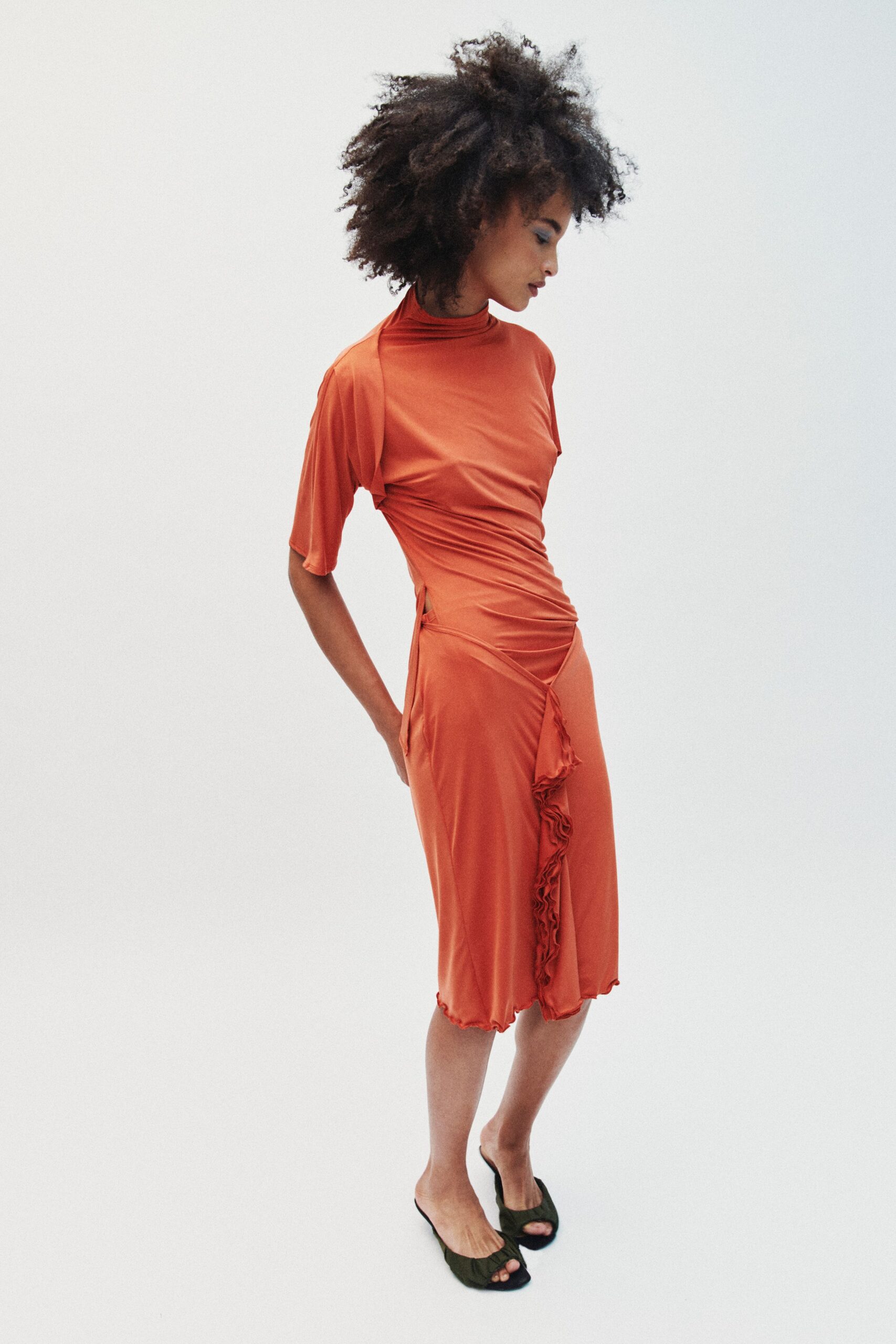 Burnt orange jersey dress, one sleeve hanging cape-like off the shoulder. Ruched through the waist with a narrow hip cutout. Black open-toe mules with gathered fabric across the vamp. Walking home barefoot-minded after dancing on a sticky roof terrace.